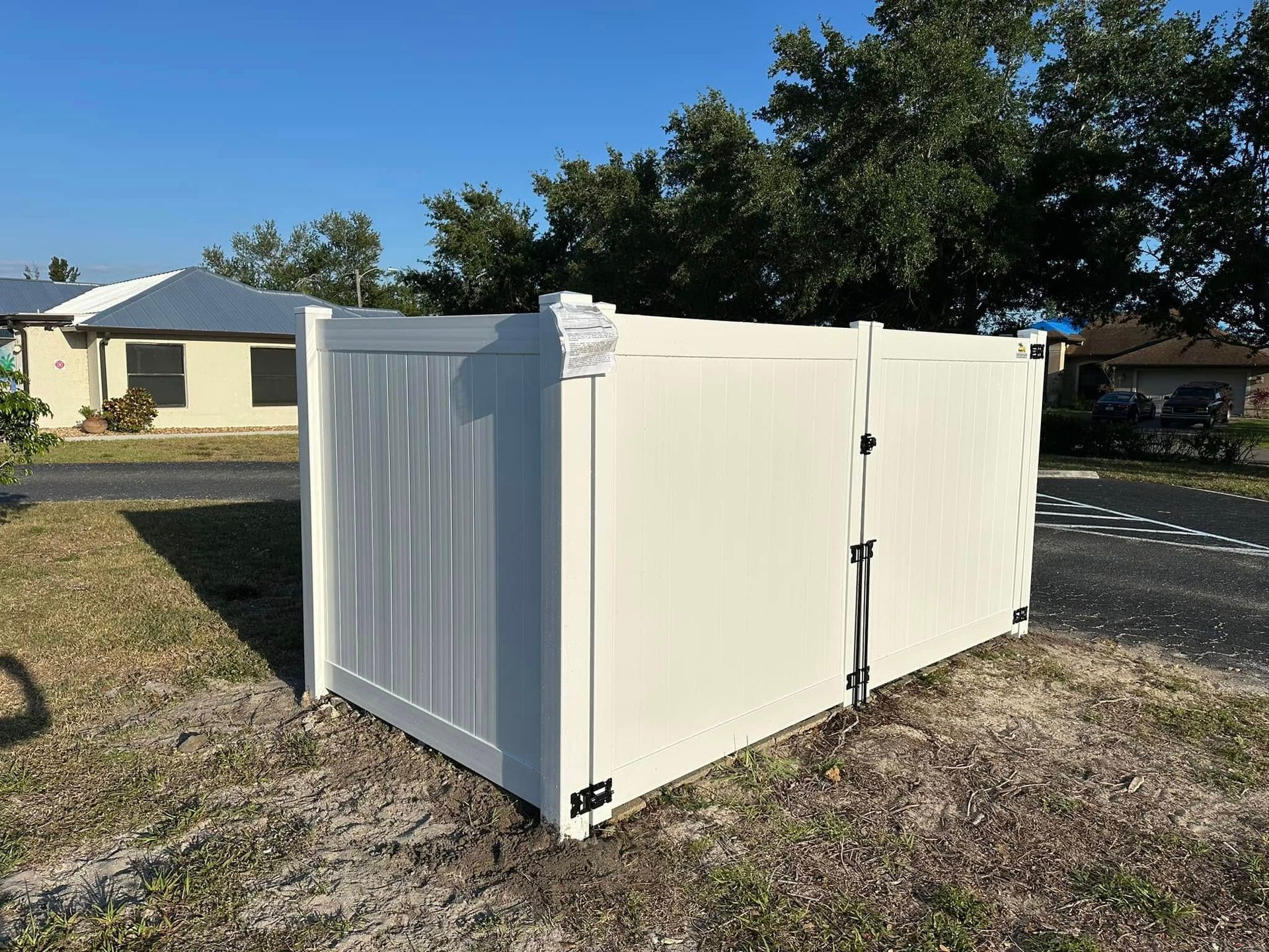 White vinyl fence with a gate on a grassy area. Houses and trees are in the background.