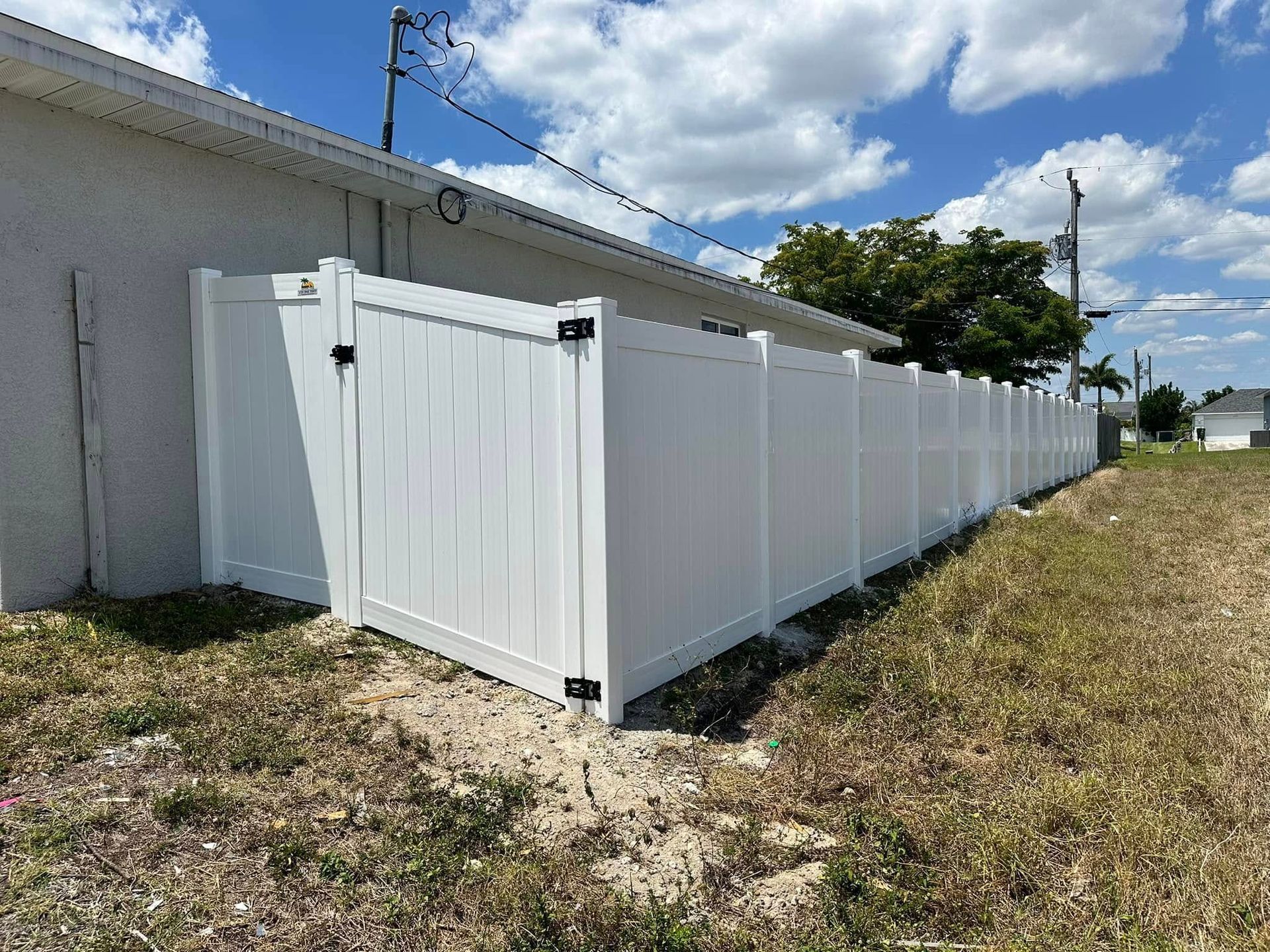 White vinyl fence with gate, extending along side of a house, under a partly cloudy blue sky.