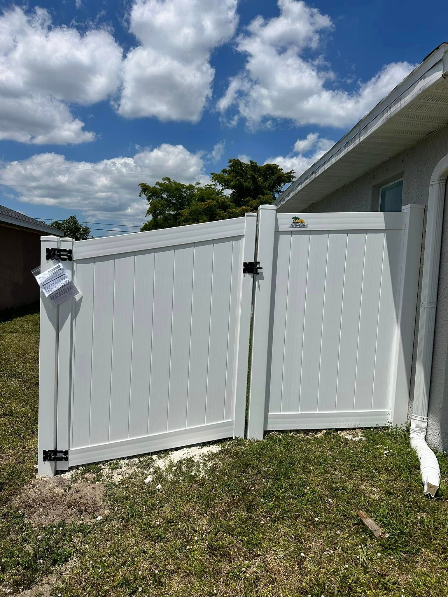 White vinyl fence with two gates, standing in grassy area against a blue sky.