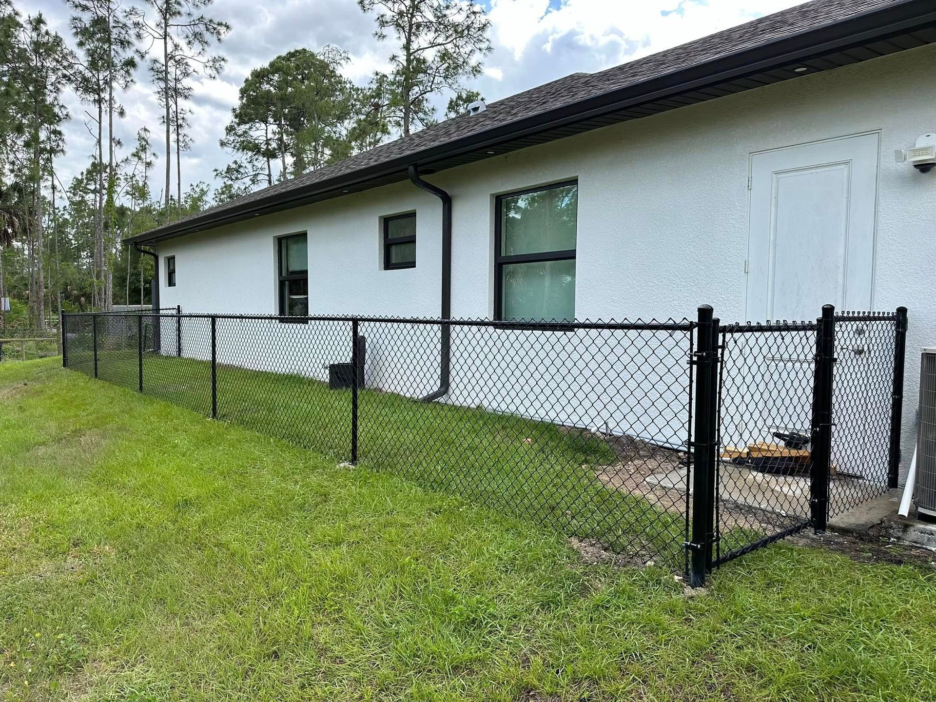 Black chain-link fence surrounds a white house with dark trim, on a grassy lawn, trees in background.