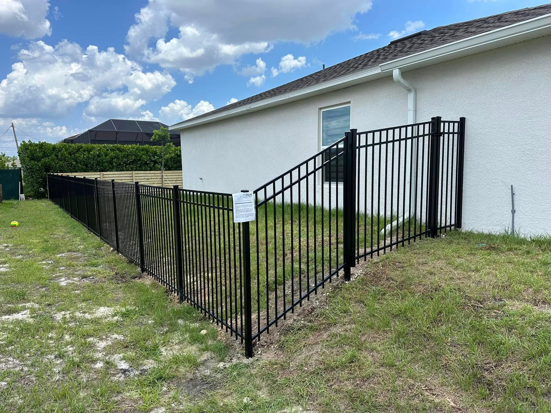 Black metal fence along a white house and grassy yard under a blue sky.