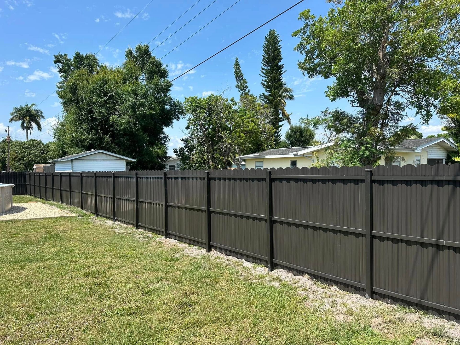 Brown privacy fence in a grassy backyard on a sunny day, houses and trees in the background.
