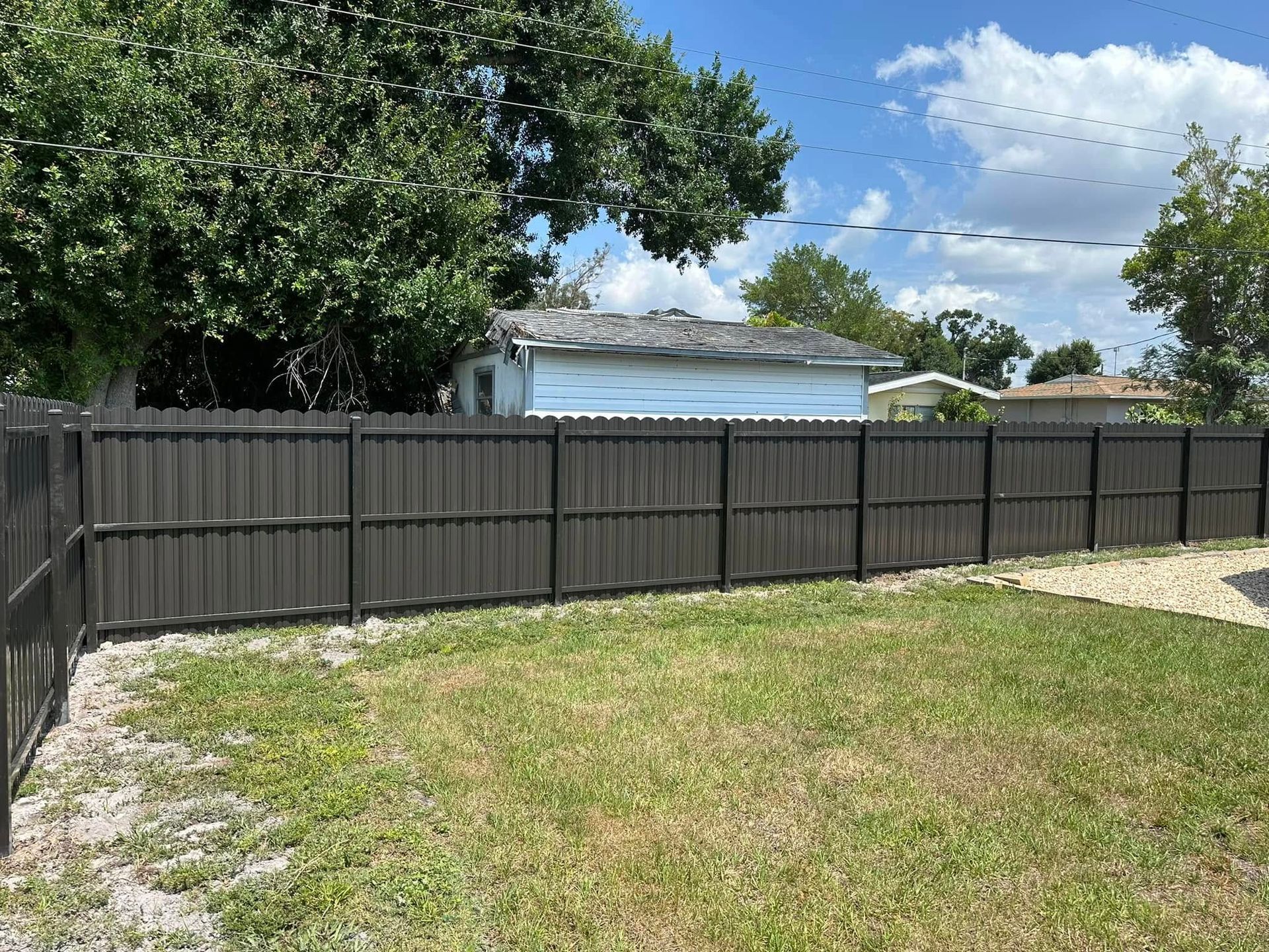 Black wooden fence in a backyard with green grass, a small house, and a blue sky.
