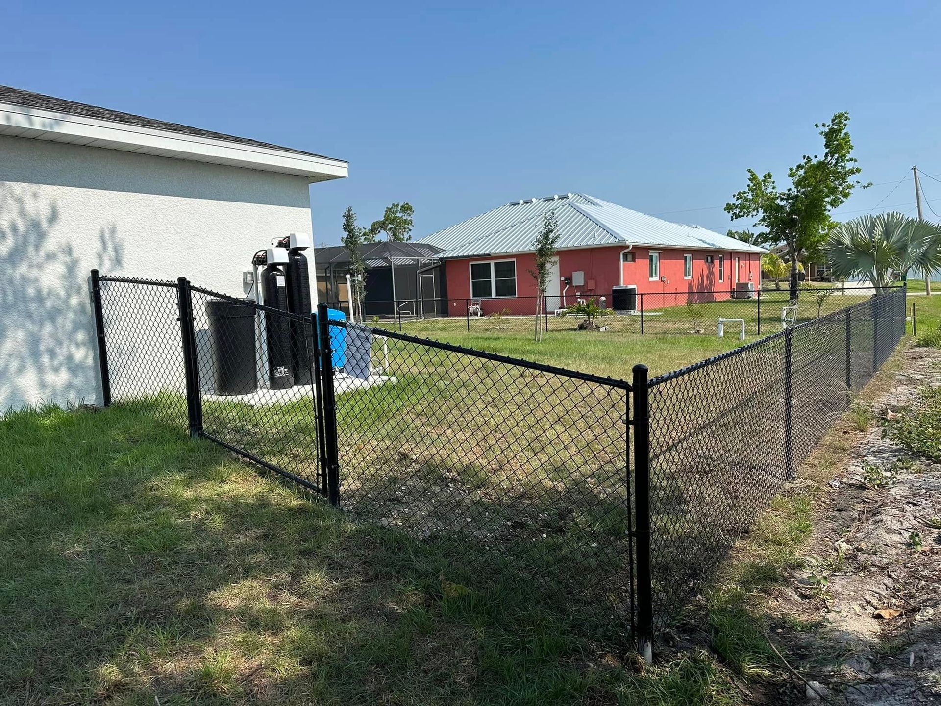 Black chain-link fence around a grassy yard, with a white building and a red house in the background under a clear blue sky.