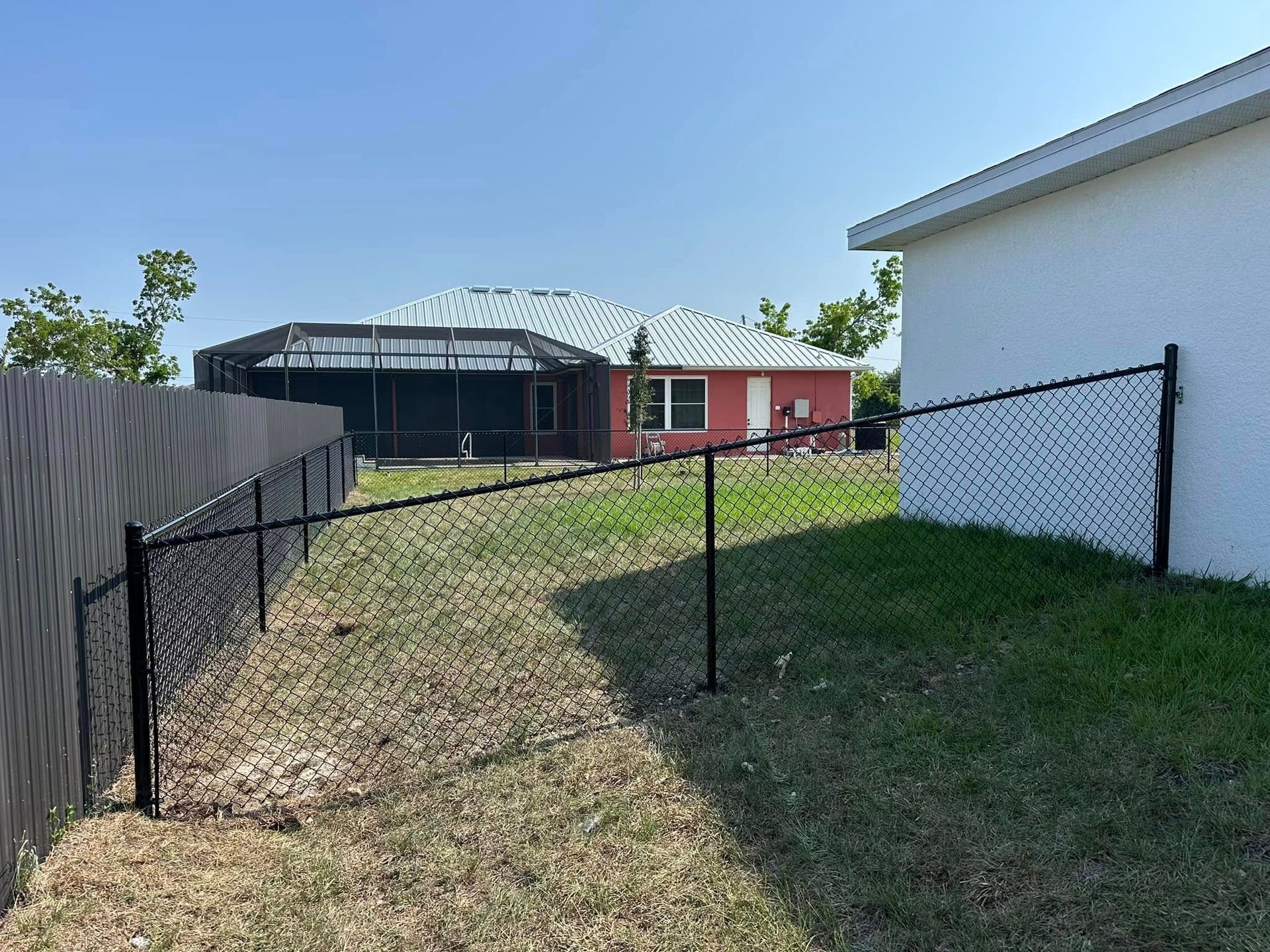 Black chain-link fence in a yard with a house in the background under a blue sky.