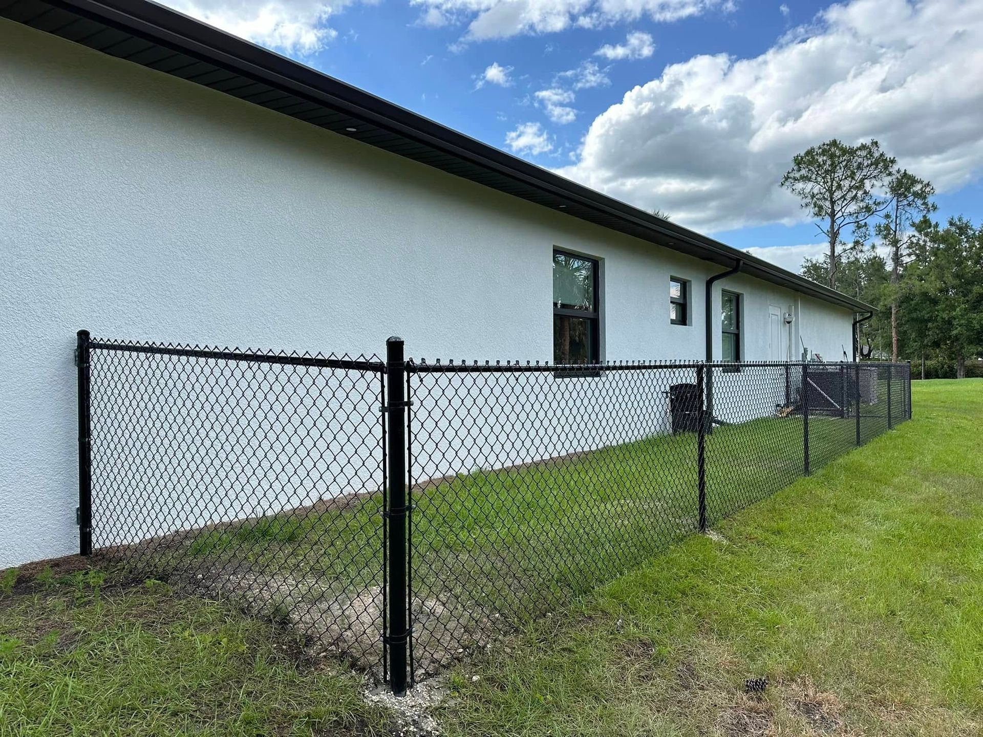 Black chain-link fence along the white stucco wall of a house; green grass and blue sky background.