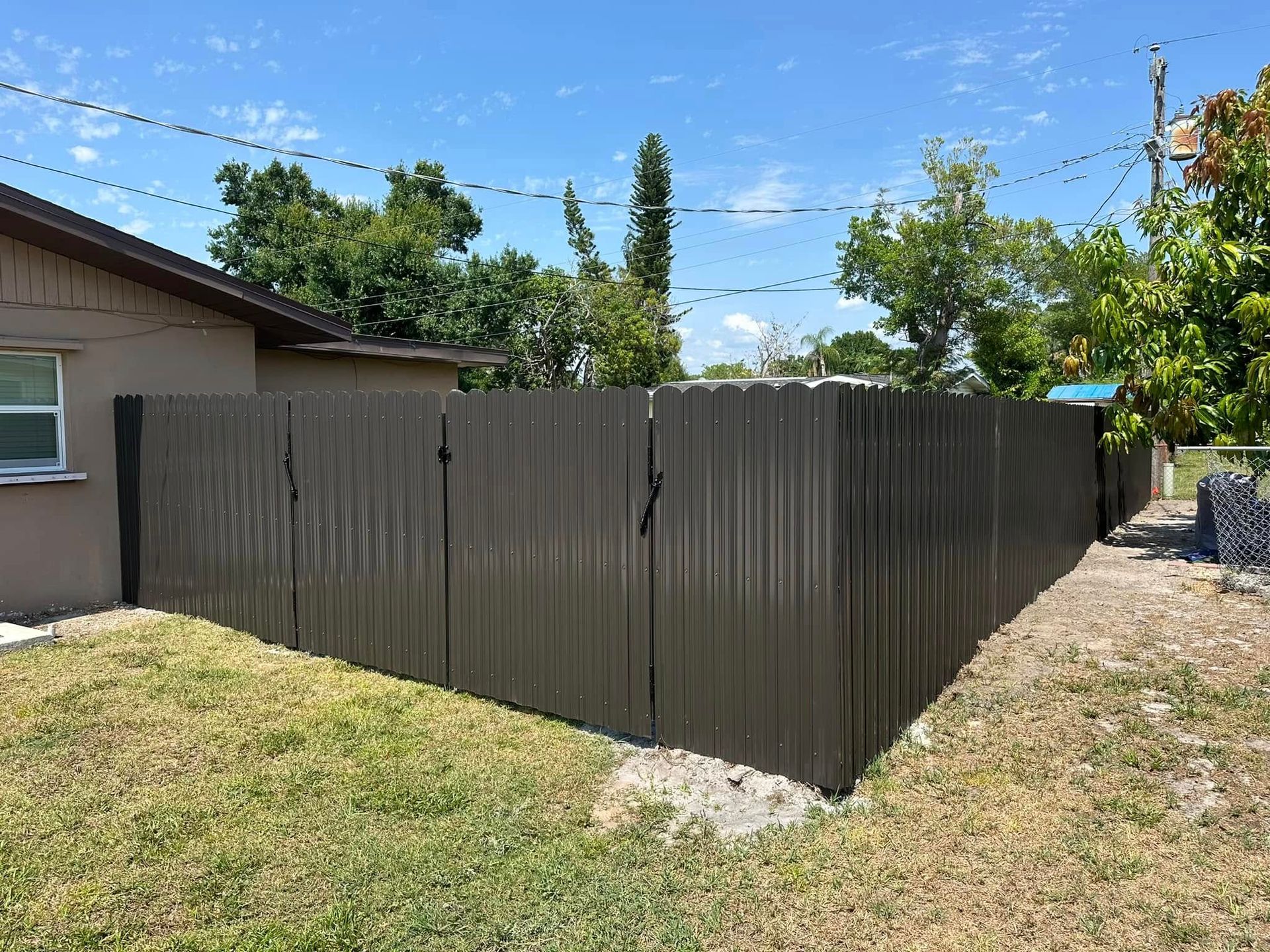 Brown corrugated metal fence in a yard, next to a house and trees, under a blue sky.