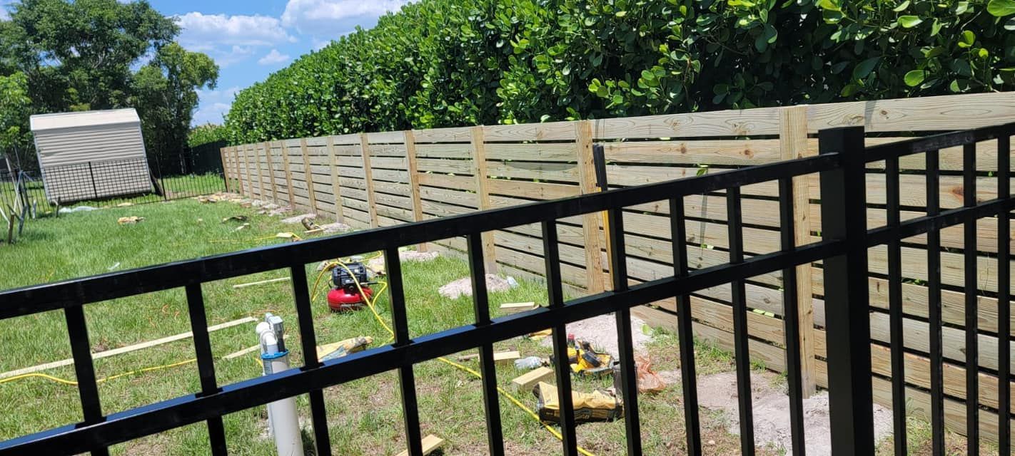 A backyard with a wooden fence, a black metal fence, and a small shed. Tools and equipment are visible on the grass.