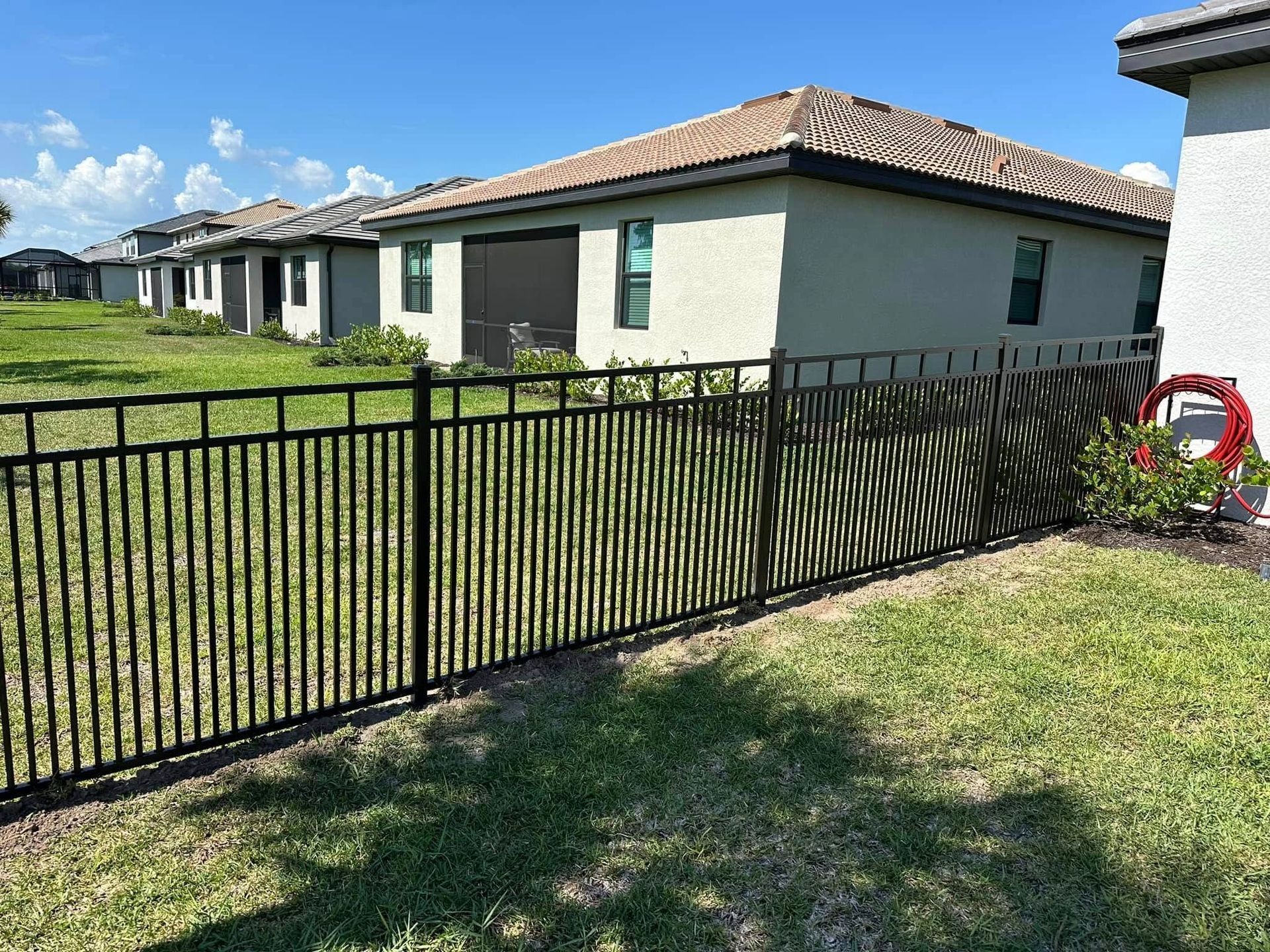 Black metal fence in front of beige houses, with green grass and blue sky.