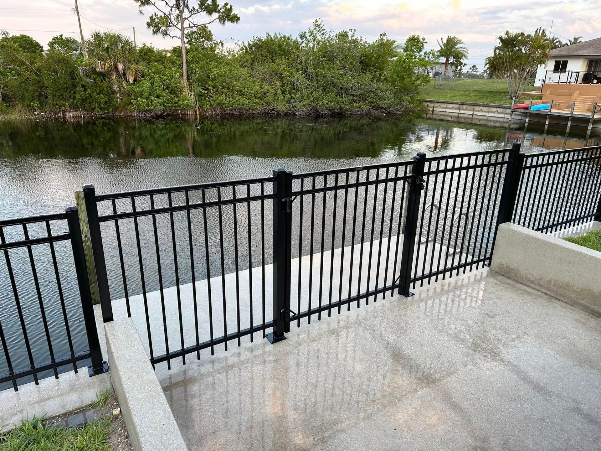 Black metal fence along a waterway, next to a concrete walkway, with lush greenery in the background.