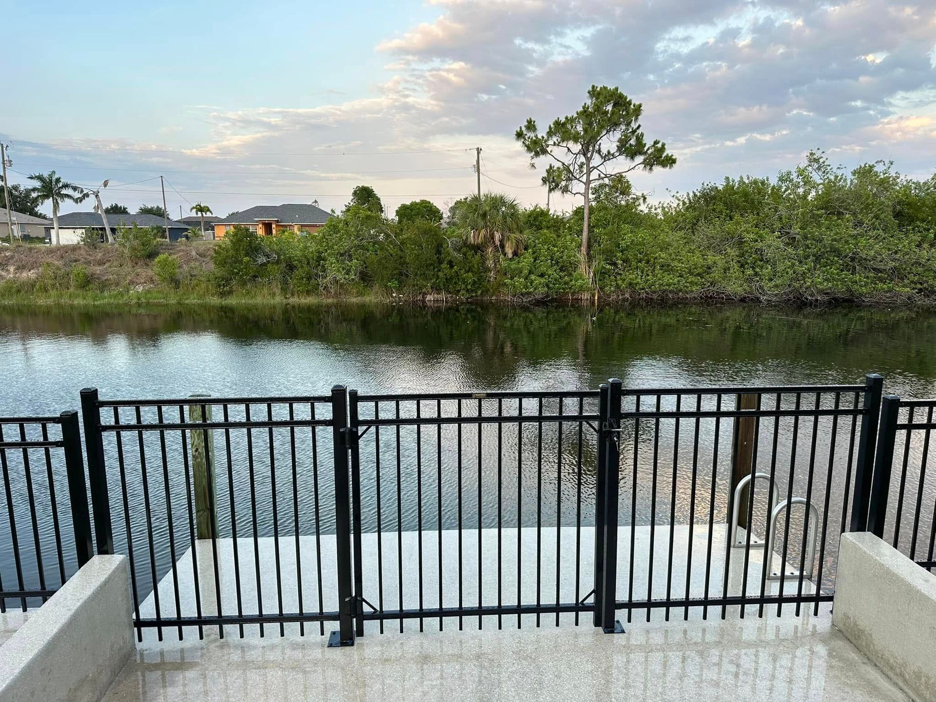 Black metal fence, dock, and canal with trees and a cloudy sky.