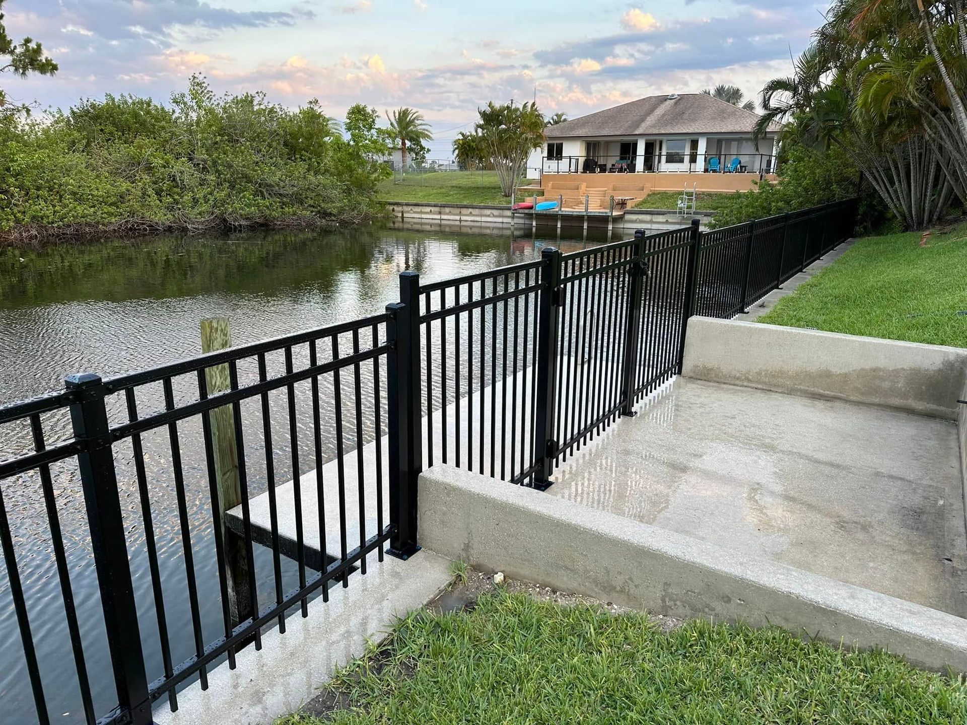 Black fence along canal, leading to a house with a dock. Green grass and trees in the background.