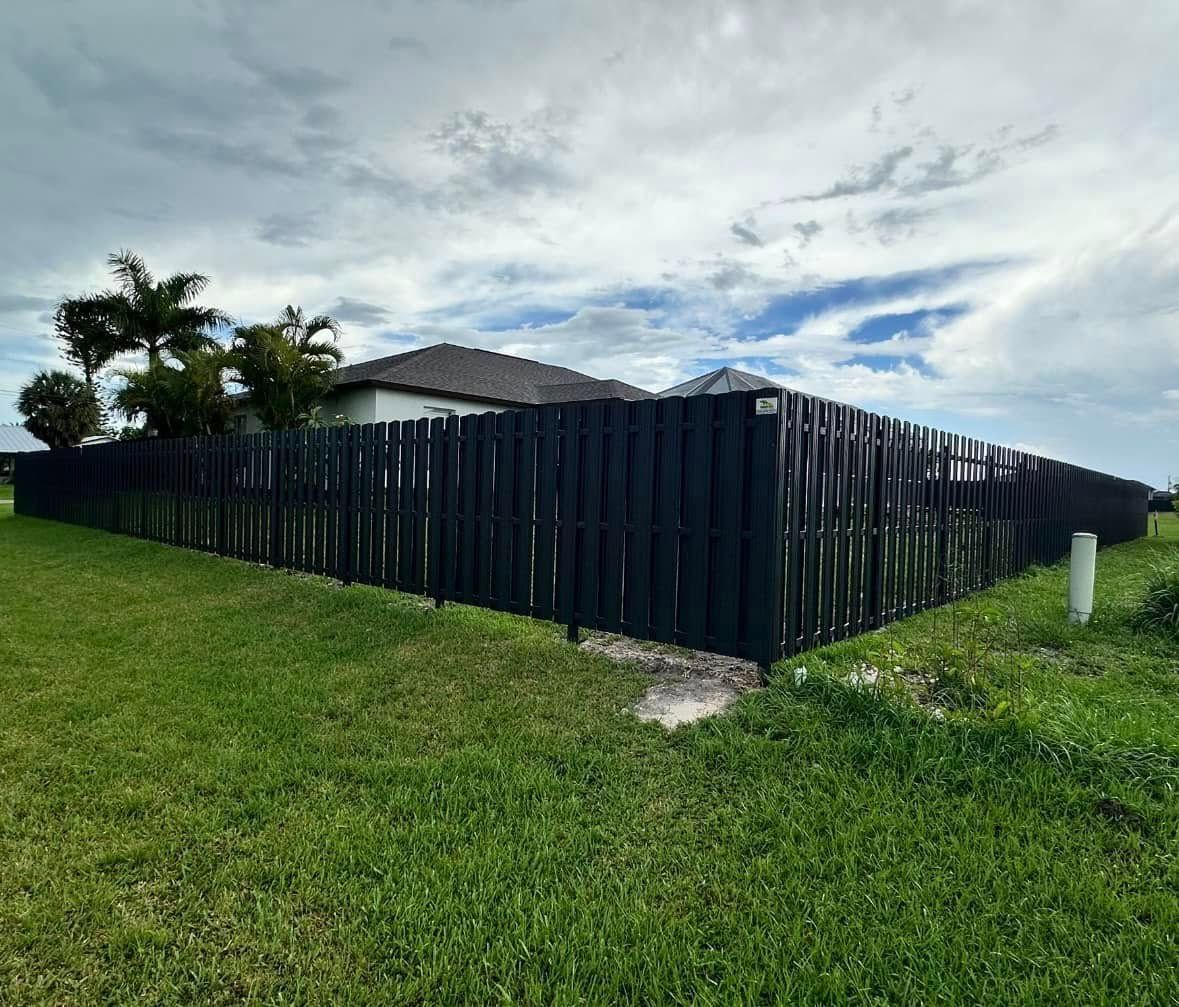 Black wooden fence around a house with green grass under a cloudy sky.