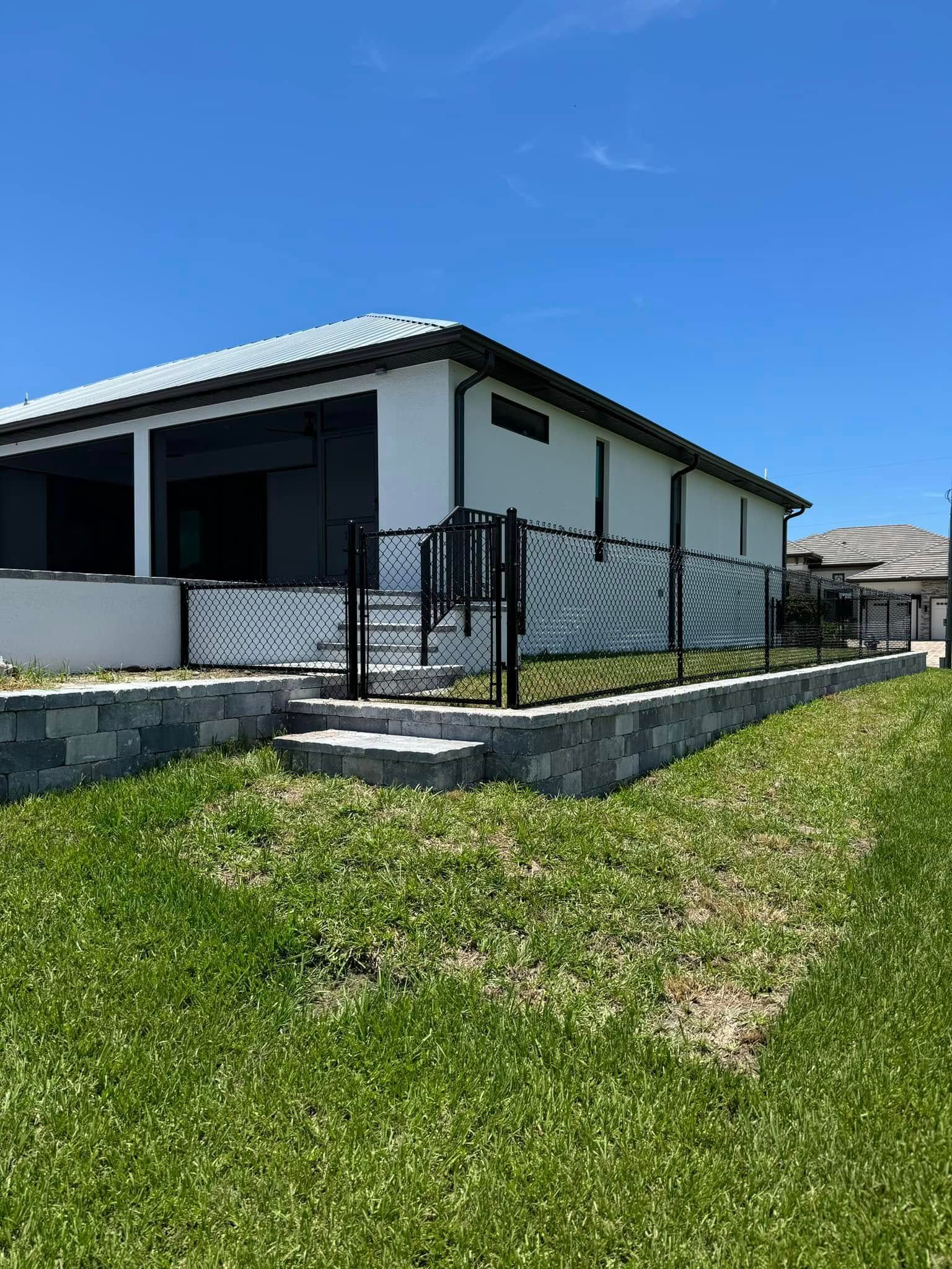 Black fence and gate around a white house with a gray retaining wall, on a grassy lawn.