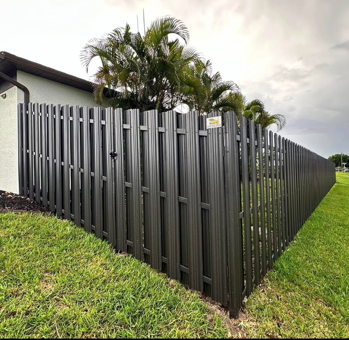 Dark gray vertical picket fence in front of a house, with green grass and palm trees in the background.