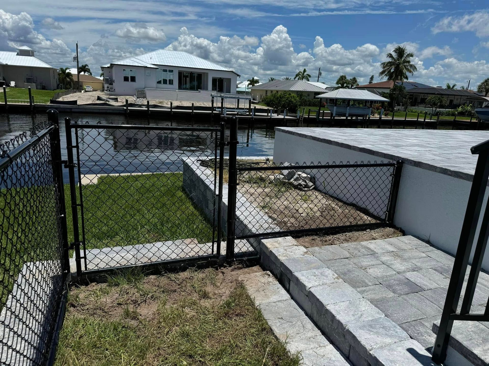 Black chain-link fence with gate on a waterway, lawn in foreground, houses and sky in background.