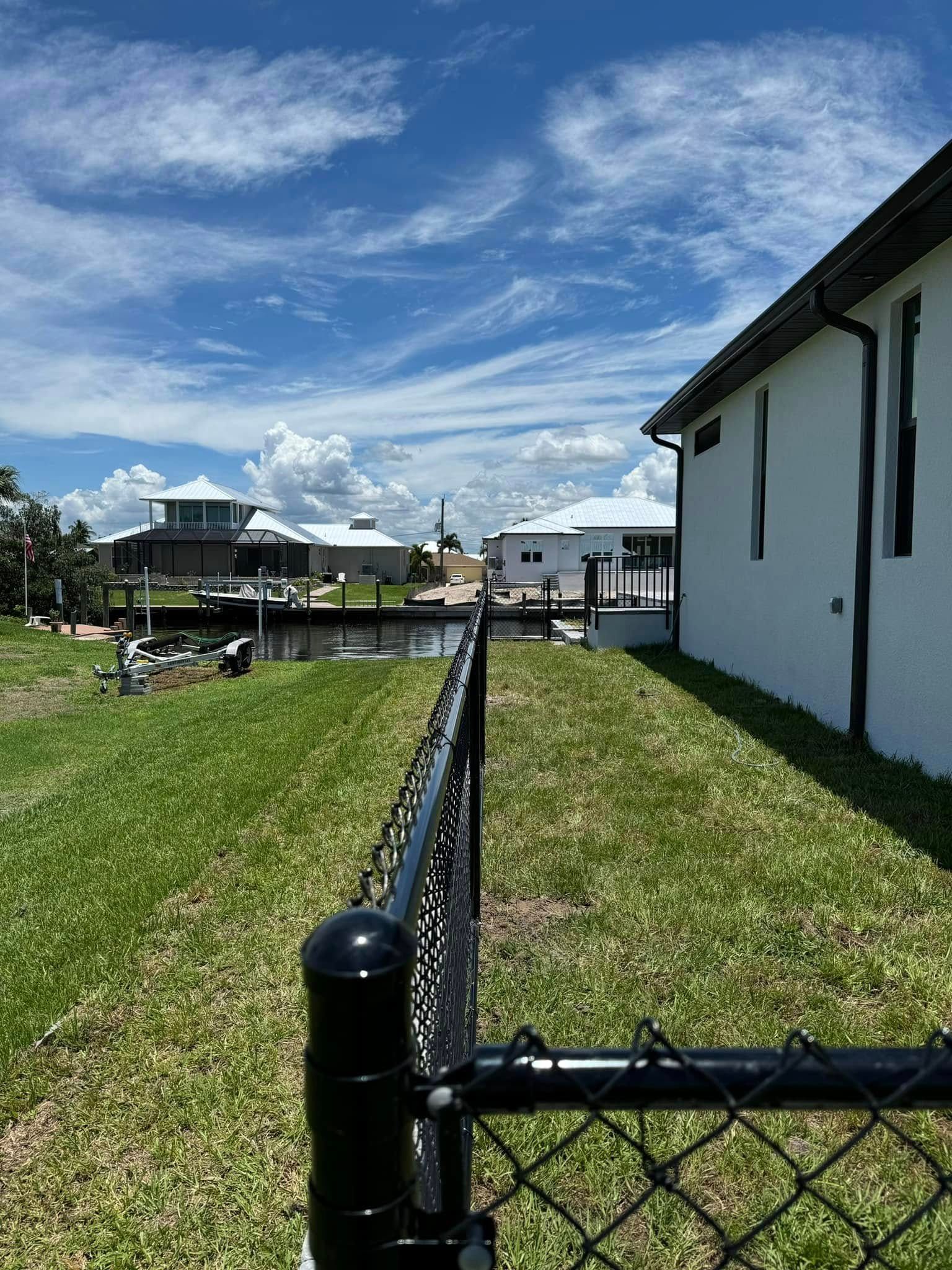 Chain-link fence beside a green lawn, leading to a canal with white houses and a cloudy blue sky.