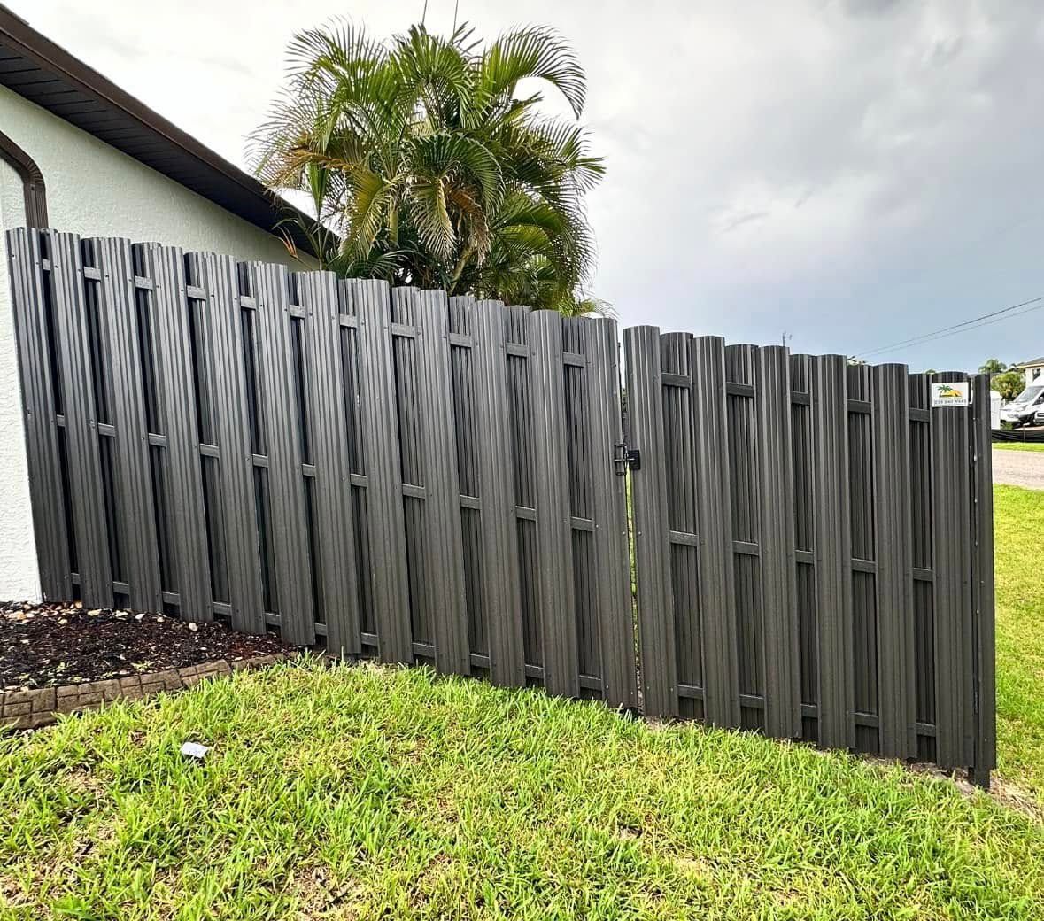 Gray textured fence along a lawn next to a white house. Cloudy sky above.