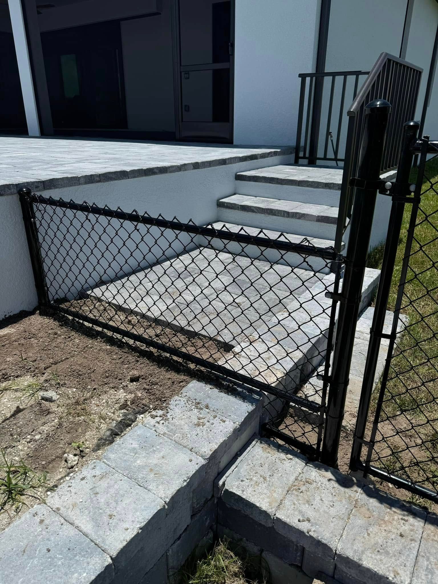 Black chain-link fence with a gate blocks access to a patio with stairs leading to a house.