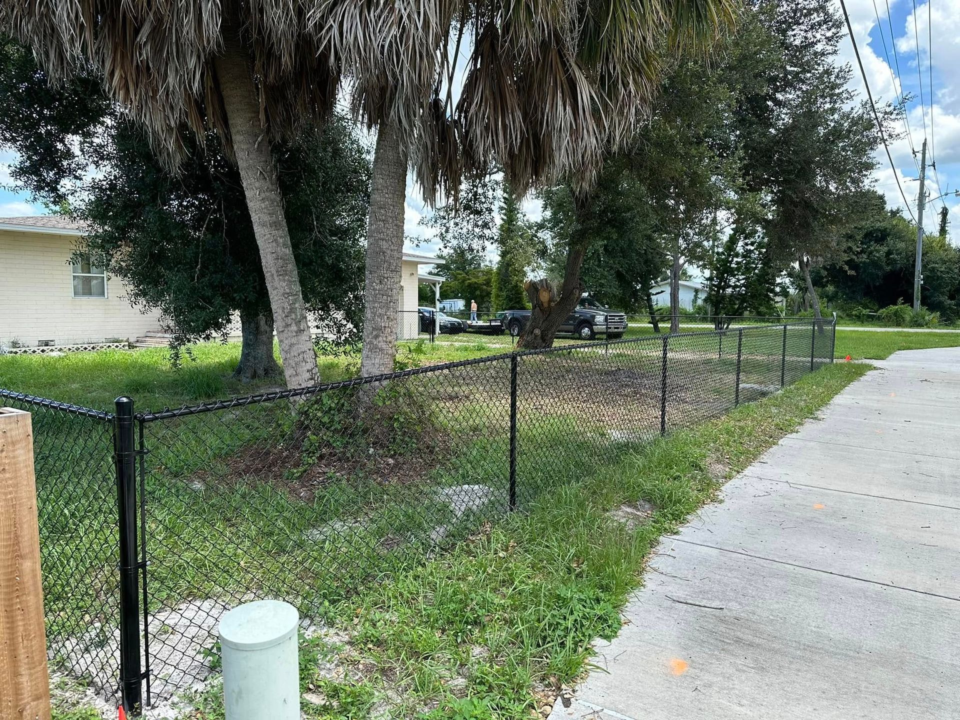 Black chain-link fence encloses a grassy yard with trees. A sidewalk and a house are visible on the right and left.