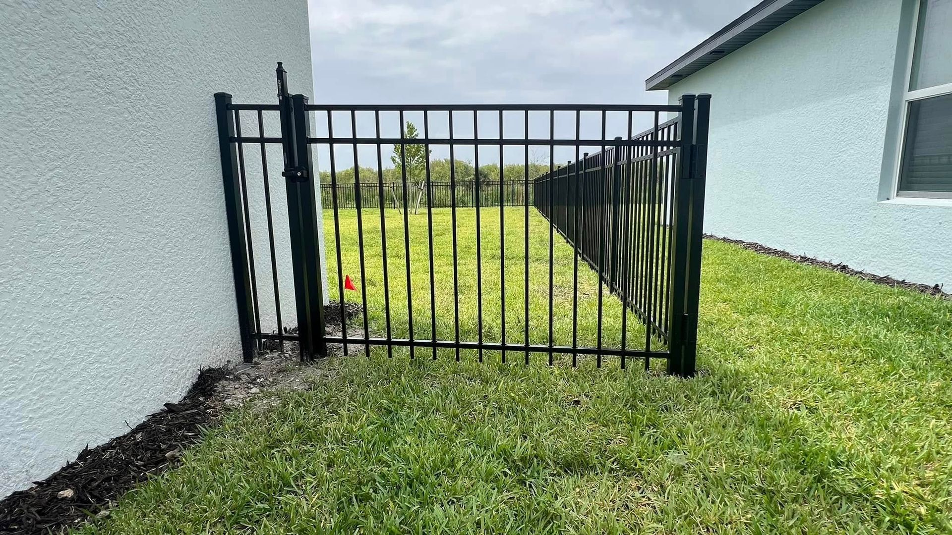 Black metal fence between two houses, enclosing a grassy area, under a cloudy sky.