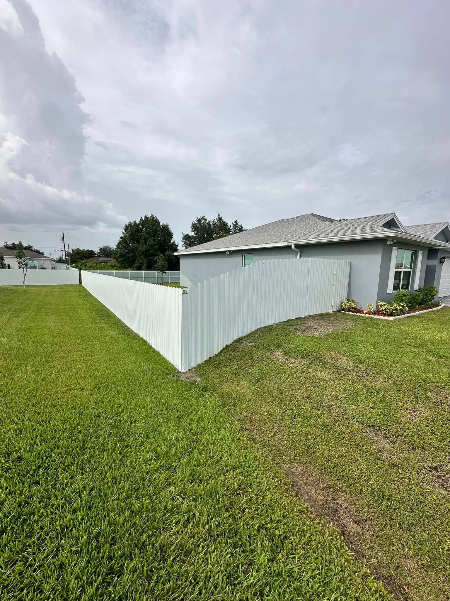 Green lawn beside a painted wooden fence, bordering a light-colored house under a cloudy sky.