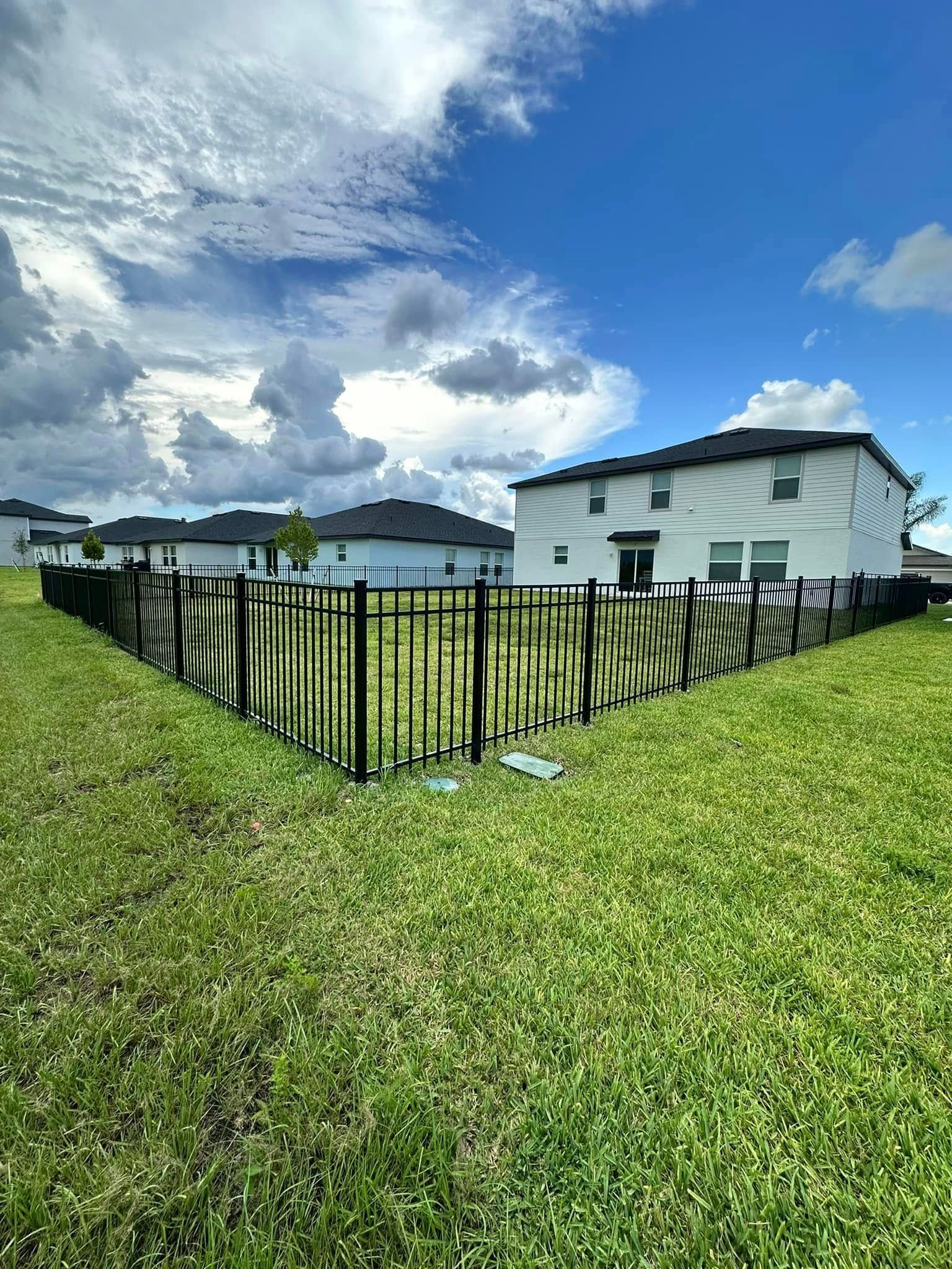 Black fence surrounding a yard of green grass; houses and blue sky in the background.