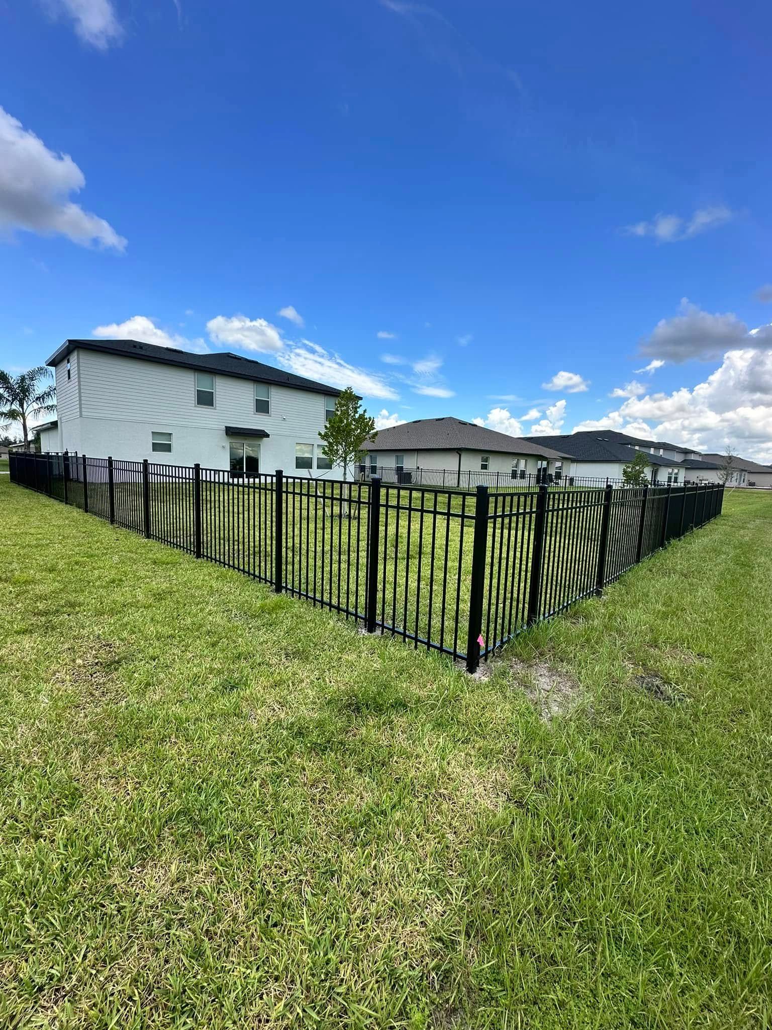 Black metal fence surrounds a grassy backyard with a two-story white house and blue sky in the background.