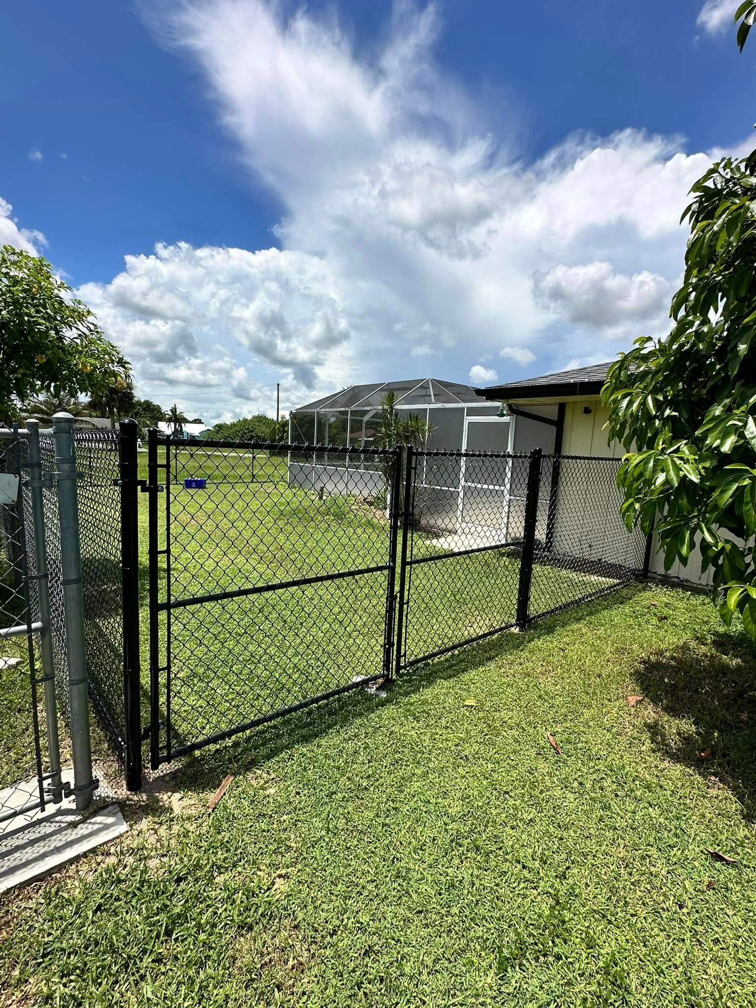 Black gate opens onto a grassy yard, leading to a building under a bright blue sky with clouds.