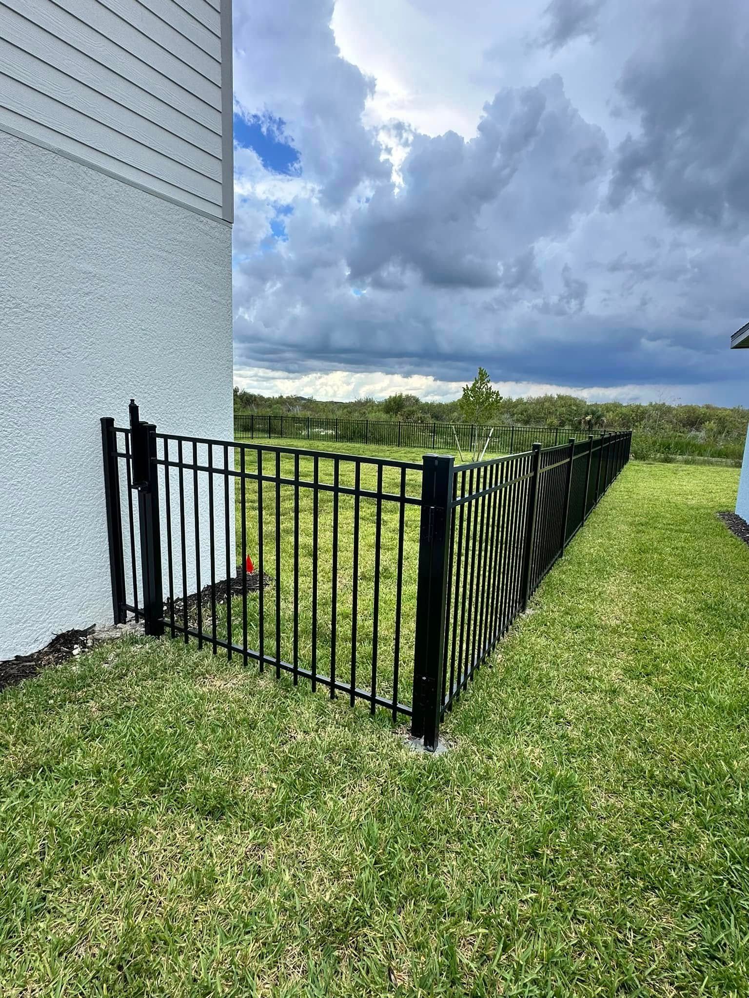 Black metal fence enclosing a grassy backyard next to a stucco house under a cloudy sky.