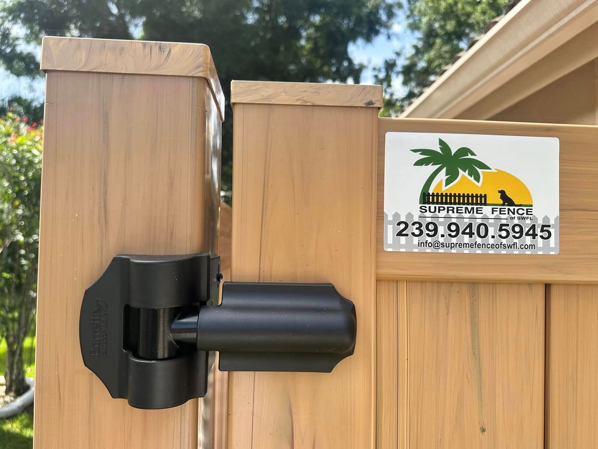 Black gate hinge on a wooden fence with a business sticker; outdoors.