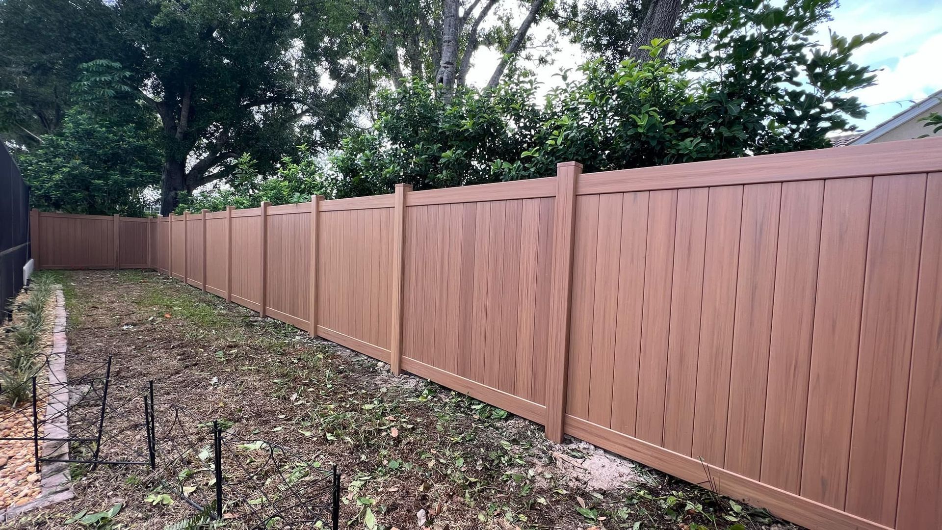 Brown wooden privacy fence surrounds a yard with sparse grass and a row of small stakes.