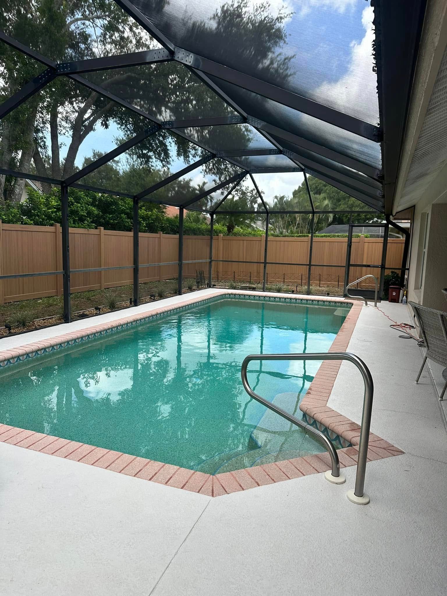 A pool enclosed by a black screen, surrounded by a light concrete patio, and a brown brick border.