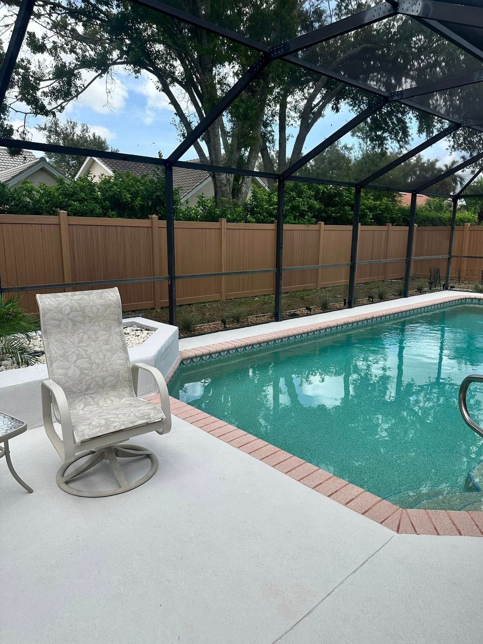 Poolside patio with a light gray chair, pool, and black screen enclosure, brown fence in the background.