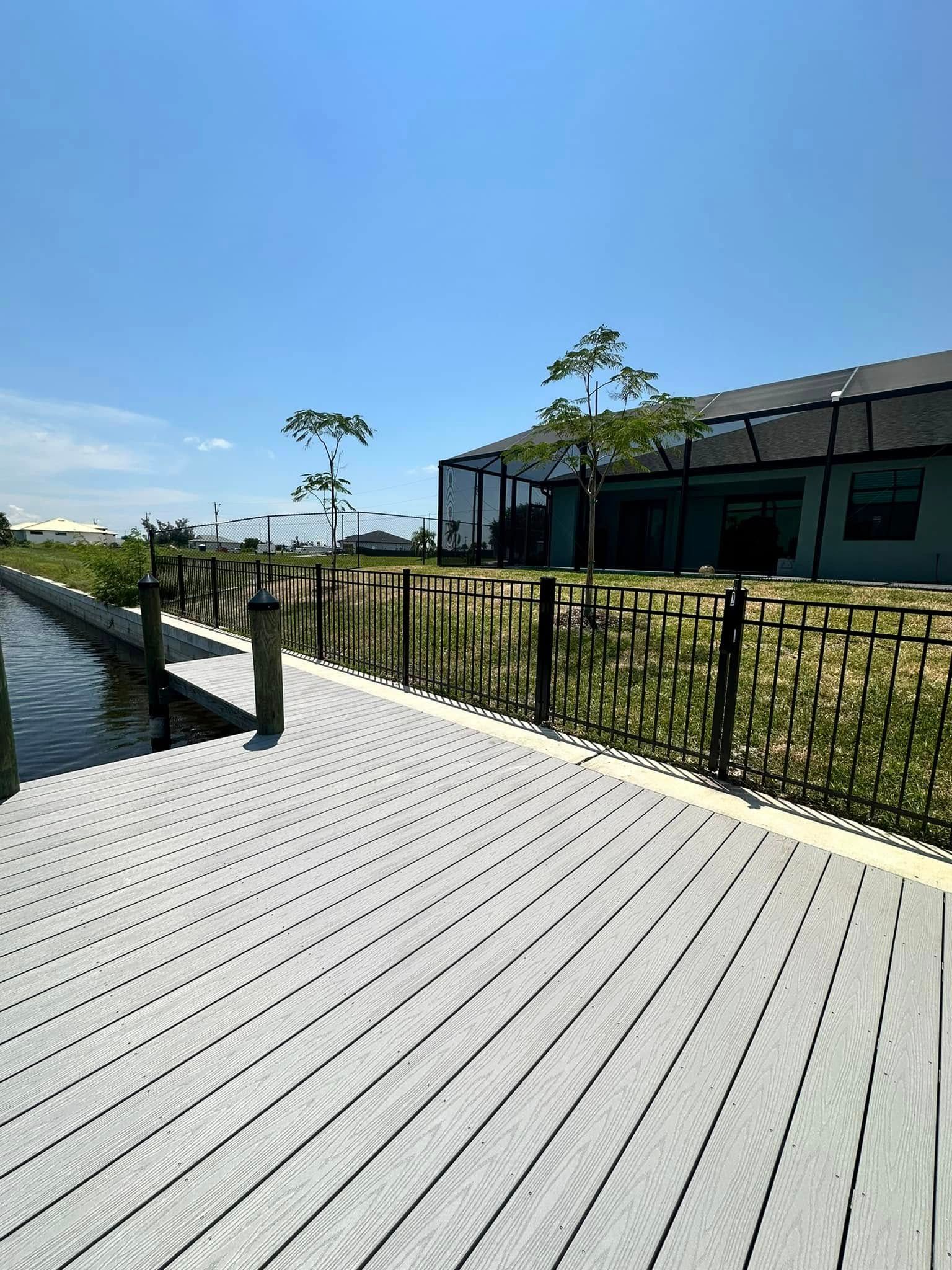 Gray composite dock alongside a canal, with a dark fence, blue sky, and house in the background.