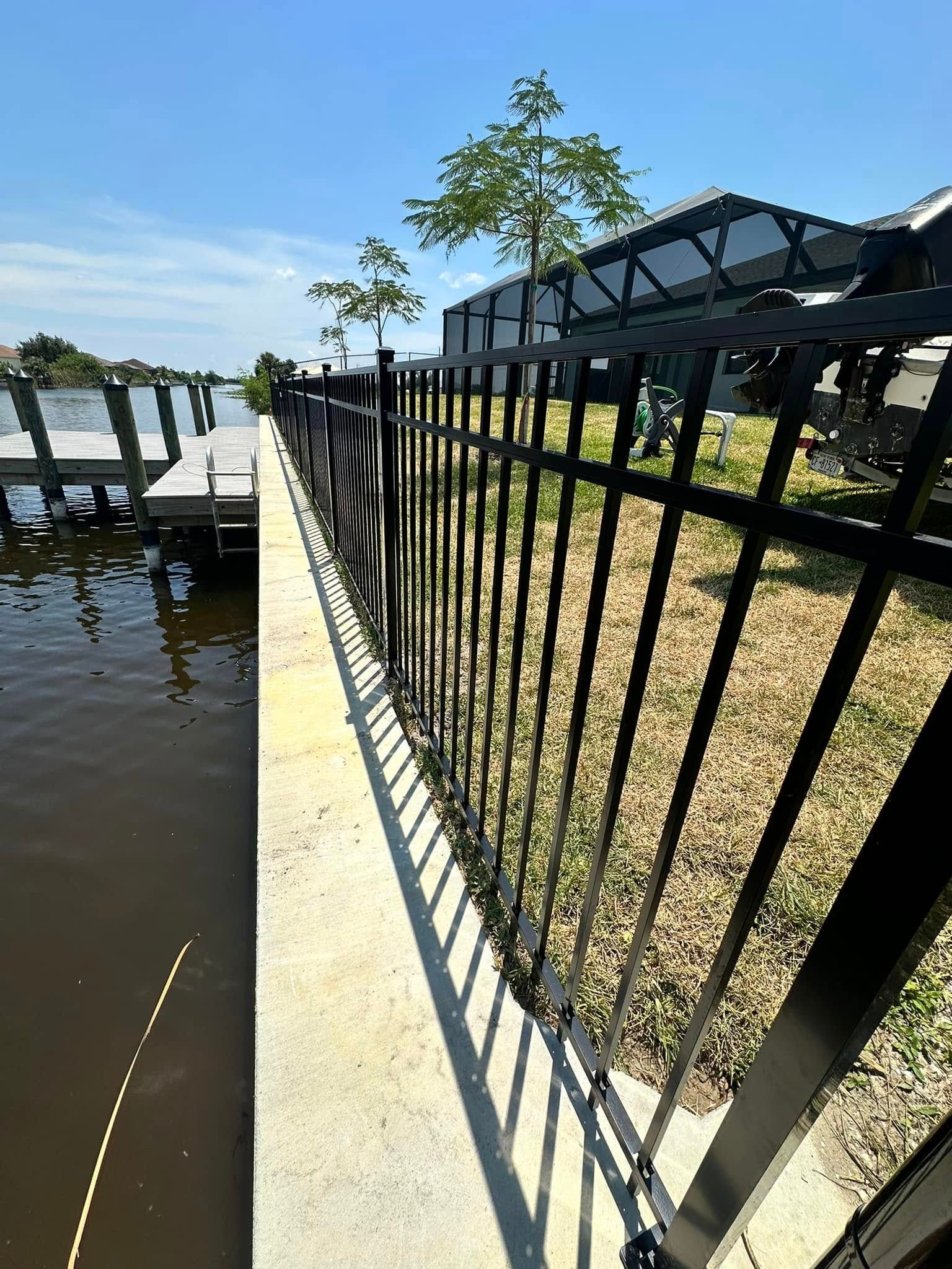 Black metal fence along a concrete walkway next to a dock on a river under a blue sky.