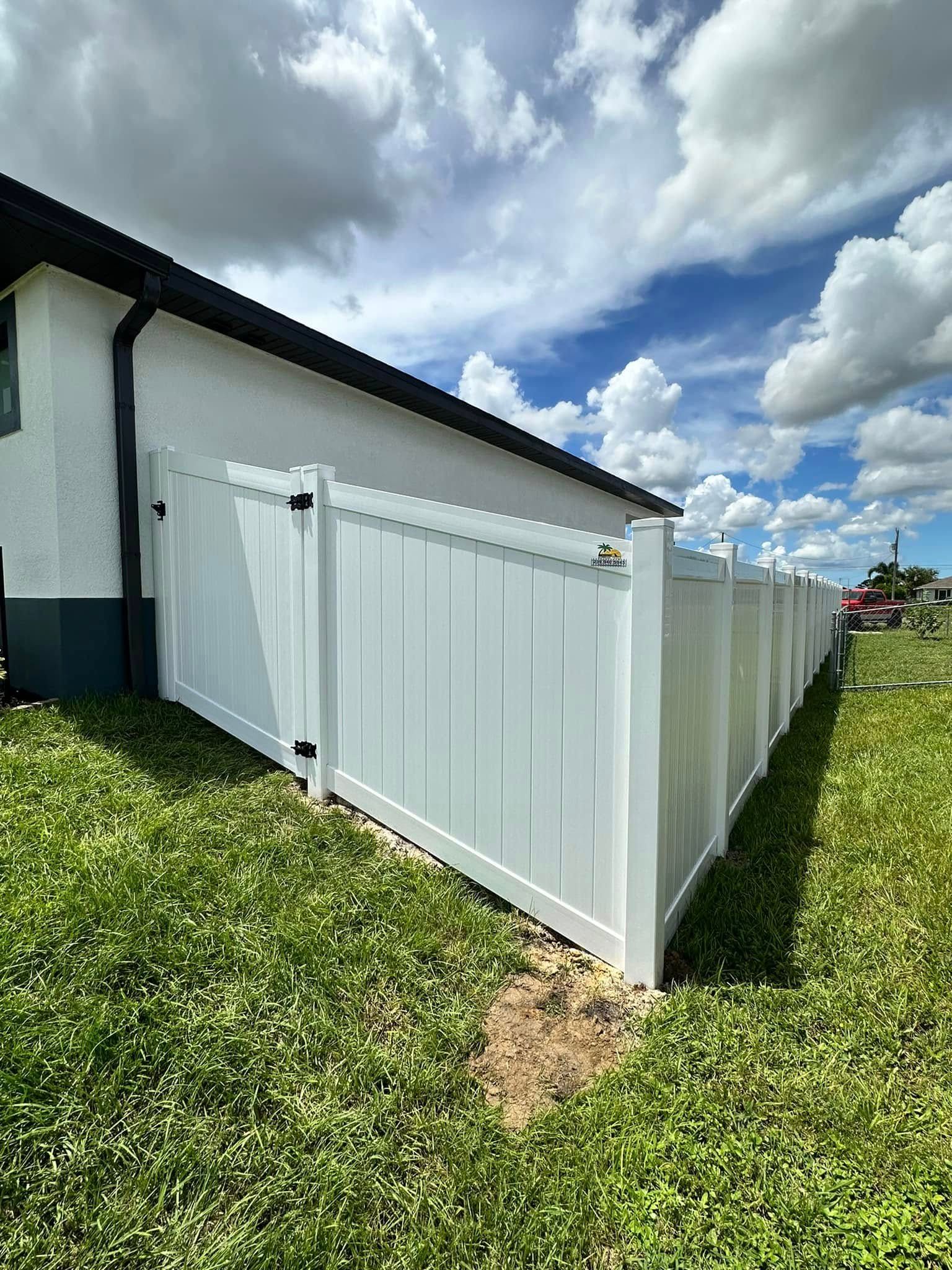 White vinyl fence surrounds a grassy yard, adjacent to a white house, under a blue sky with fluffy clouds.