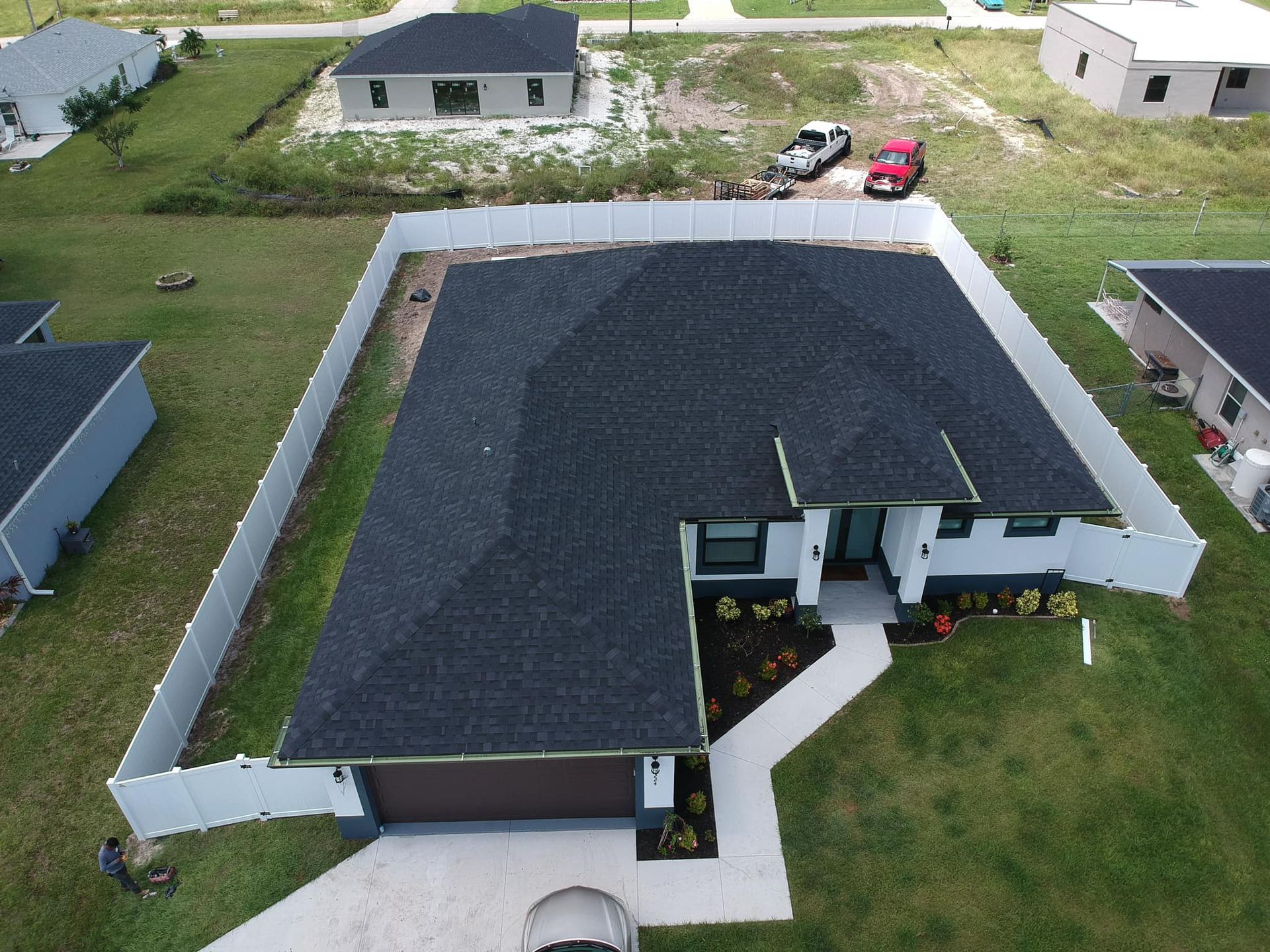 Aerial view of a house with a dark roof and white fence; vehicles and nearby houses are also visible.