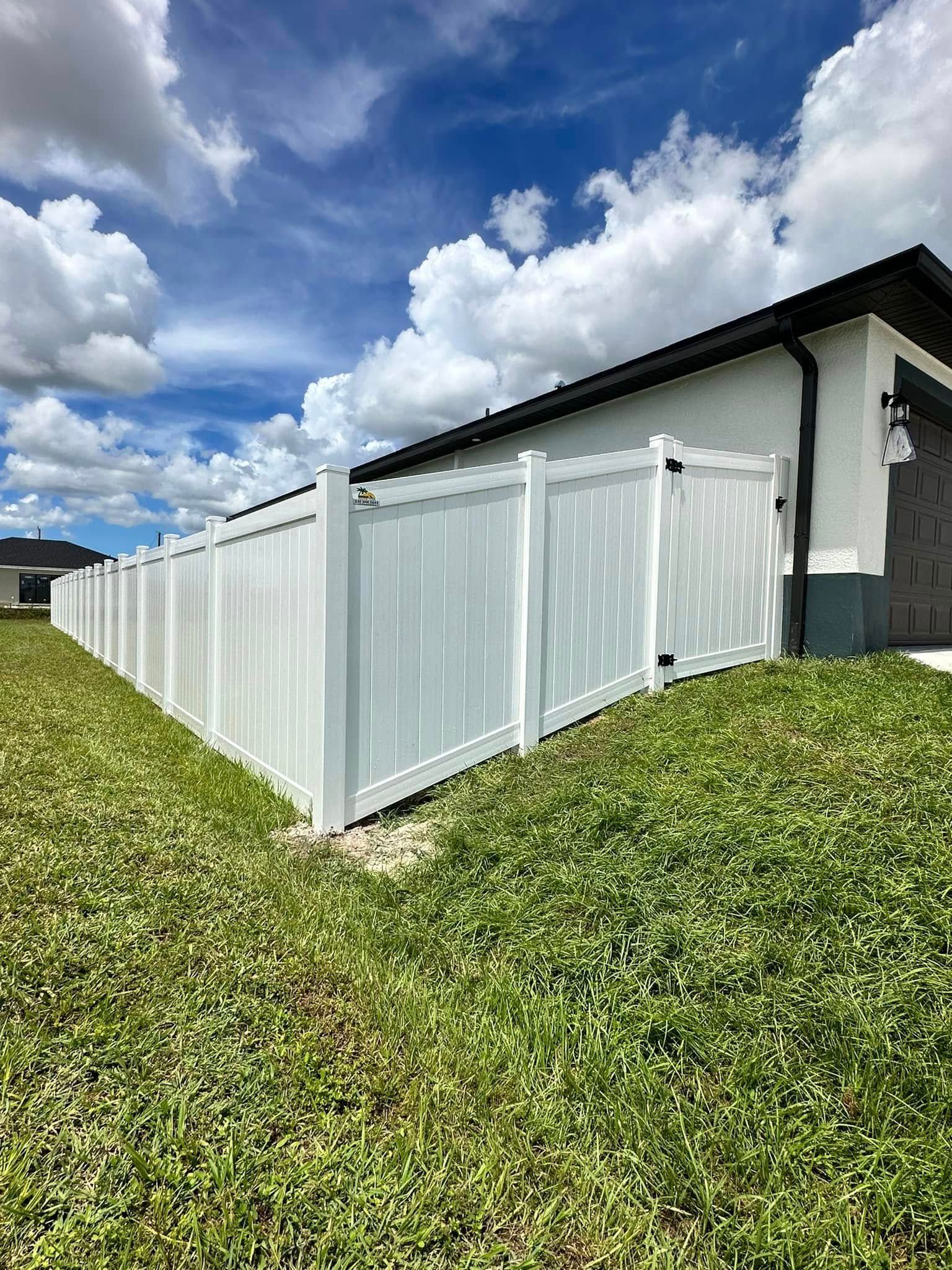 White vinyl fence alongside a house with a dark roof under a cloudy blue sky.
