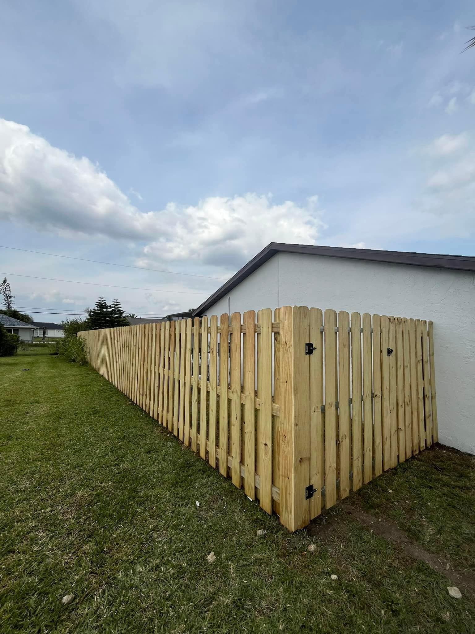 Wooden fence surrounding a grassy yard next to a white house under a cloudy sky.
