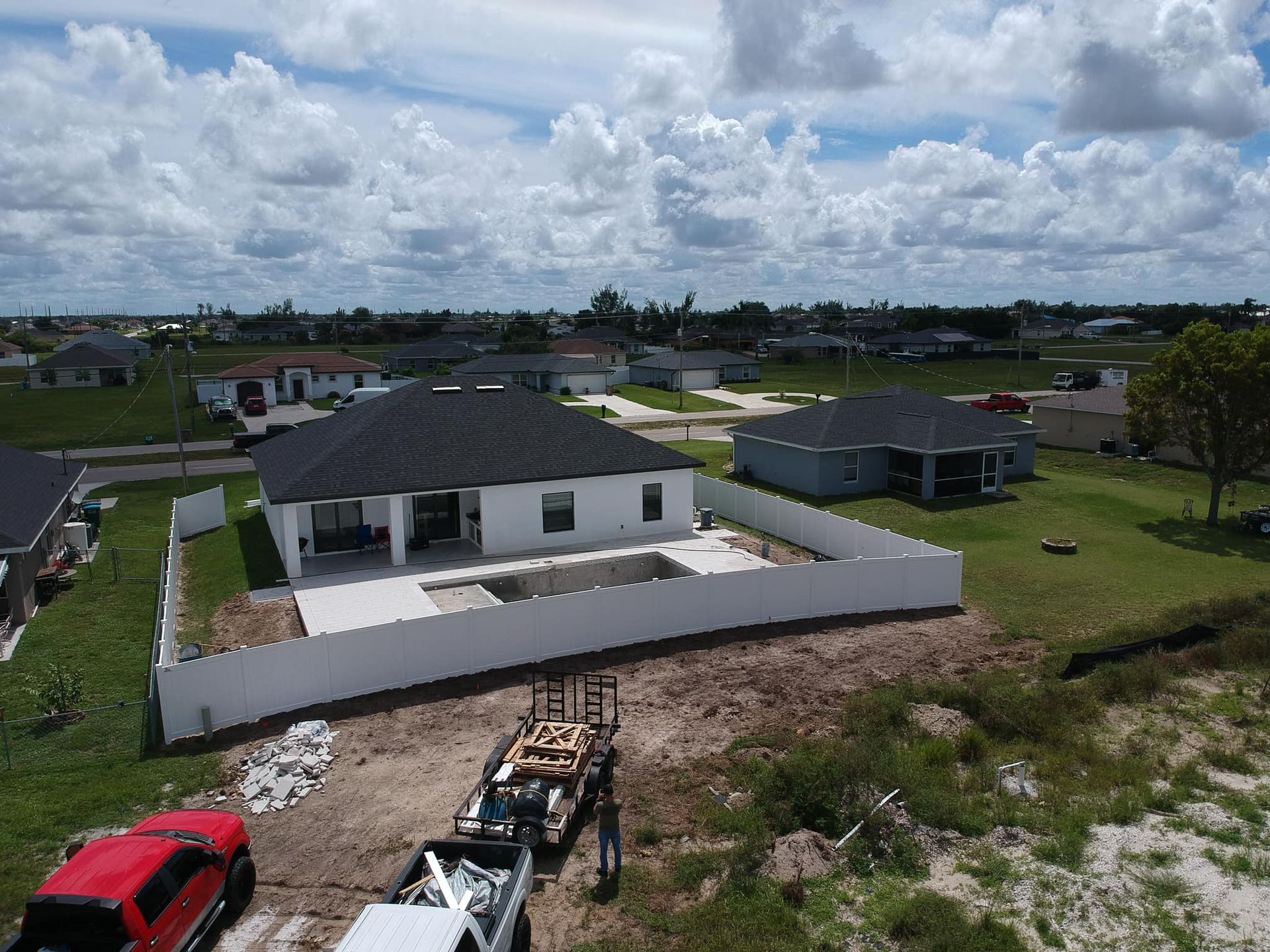 Aerial view of a new white house with a pool under construction, surrounded by a white fence in a suburban neighborhood.