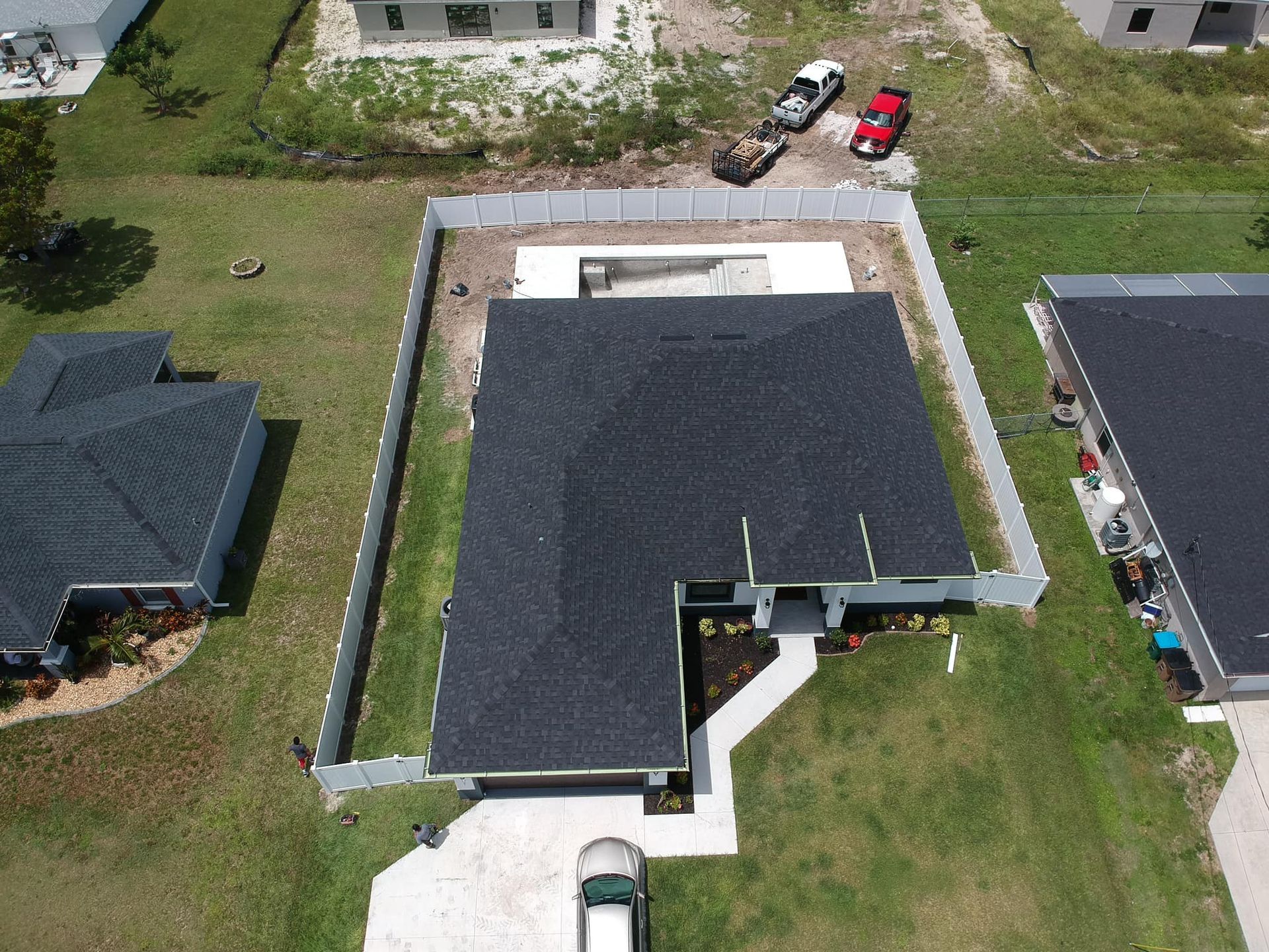 Aerial view of a house with a black roof, white fence, and a pool under construction. Cars parked nearby.