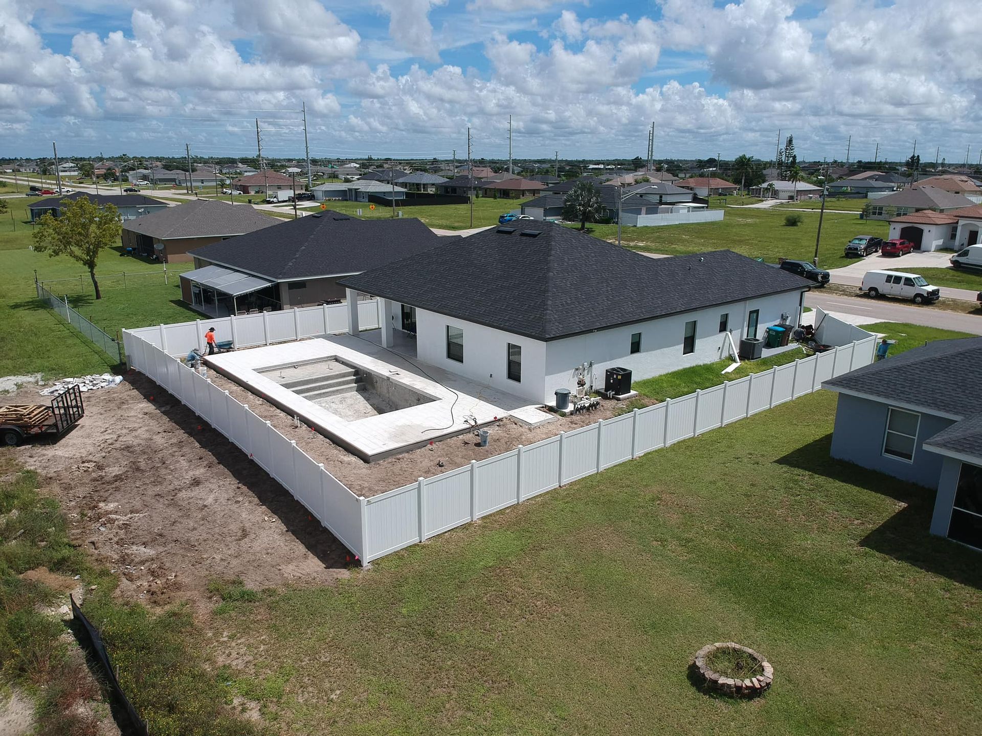 Aerial view of a white house with a pool and white fence in a suburban neighborhood under a blue sky.