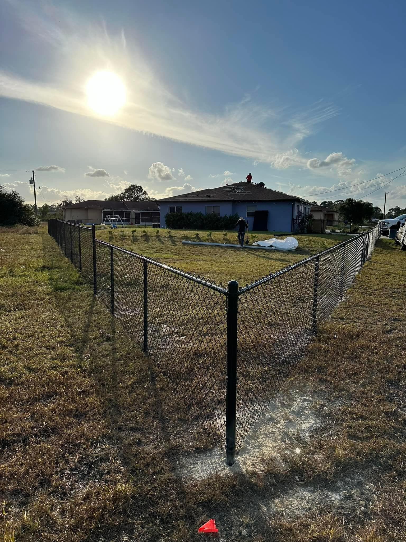 A dark fence surrounds a grassy yard in front of a blue house under a bright sun.