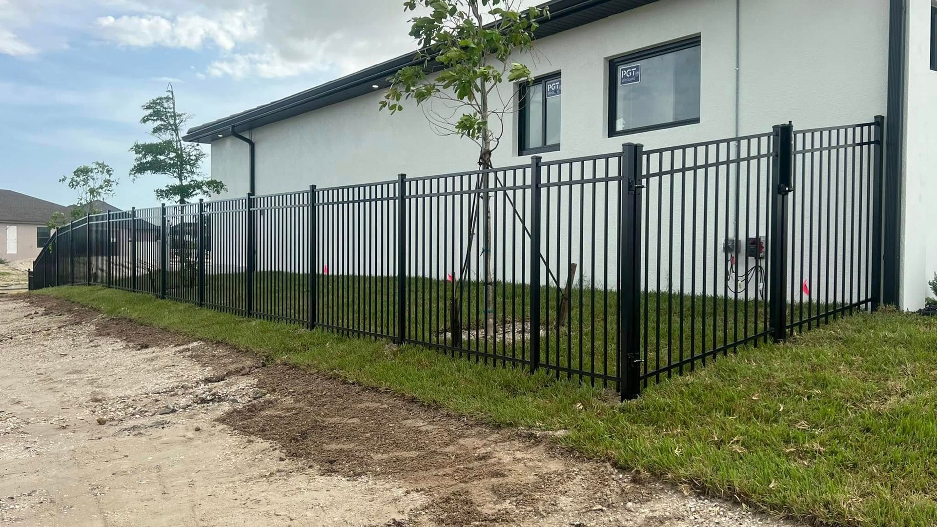 Black metal fence borders a white house on a grassy area; dirt road in the foreground.