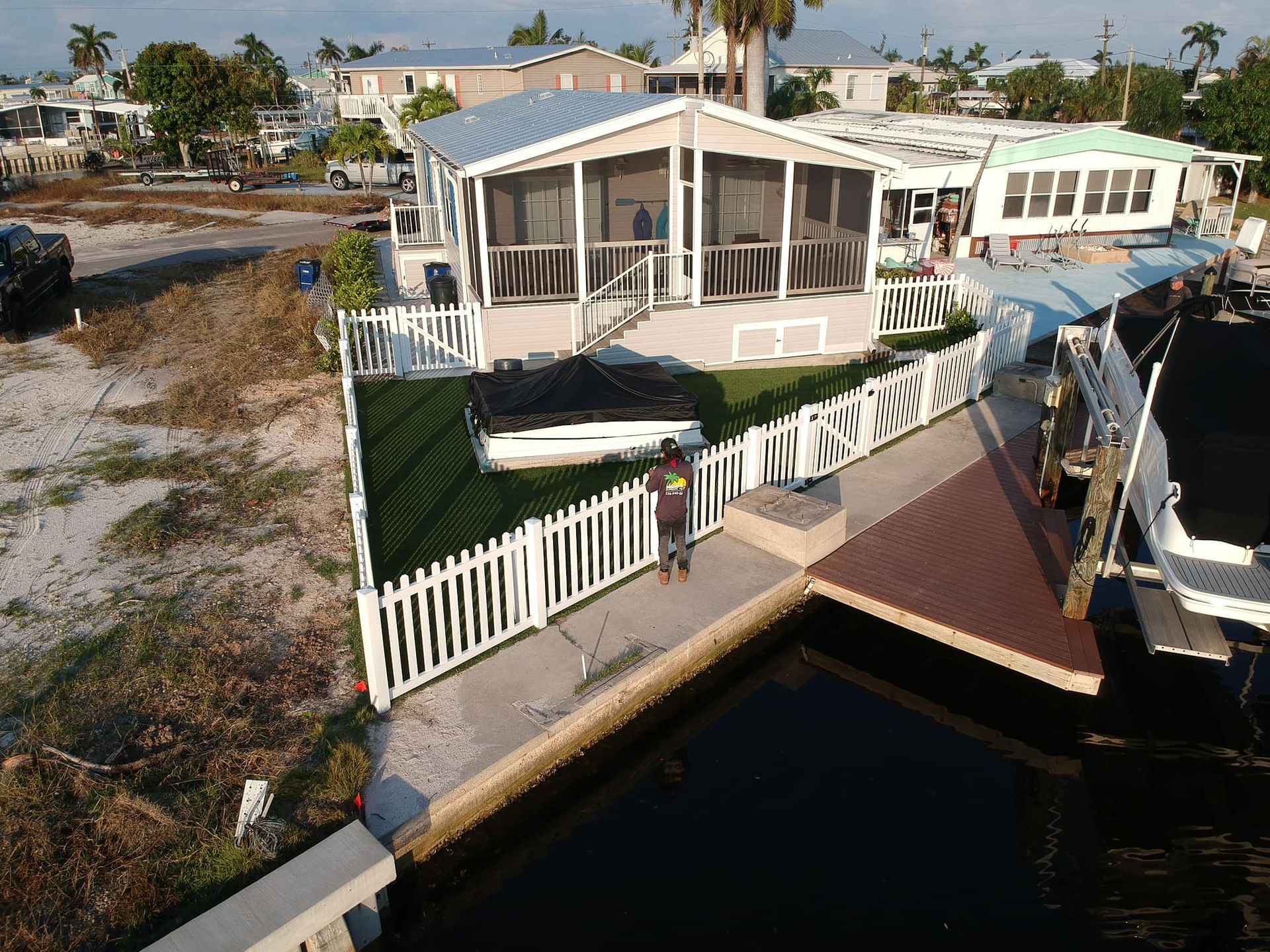 Overhead view: person fishing off a dock next to a home with a screened porch, white picket fence, and green lawn.