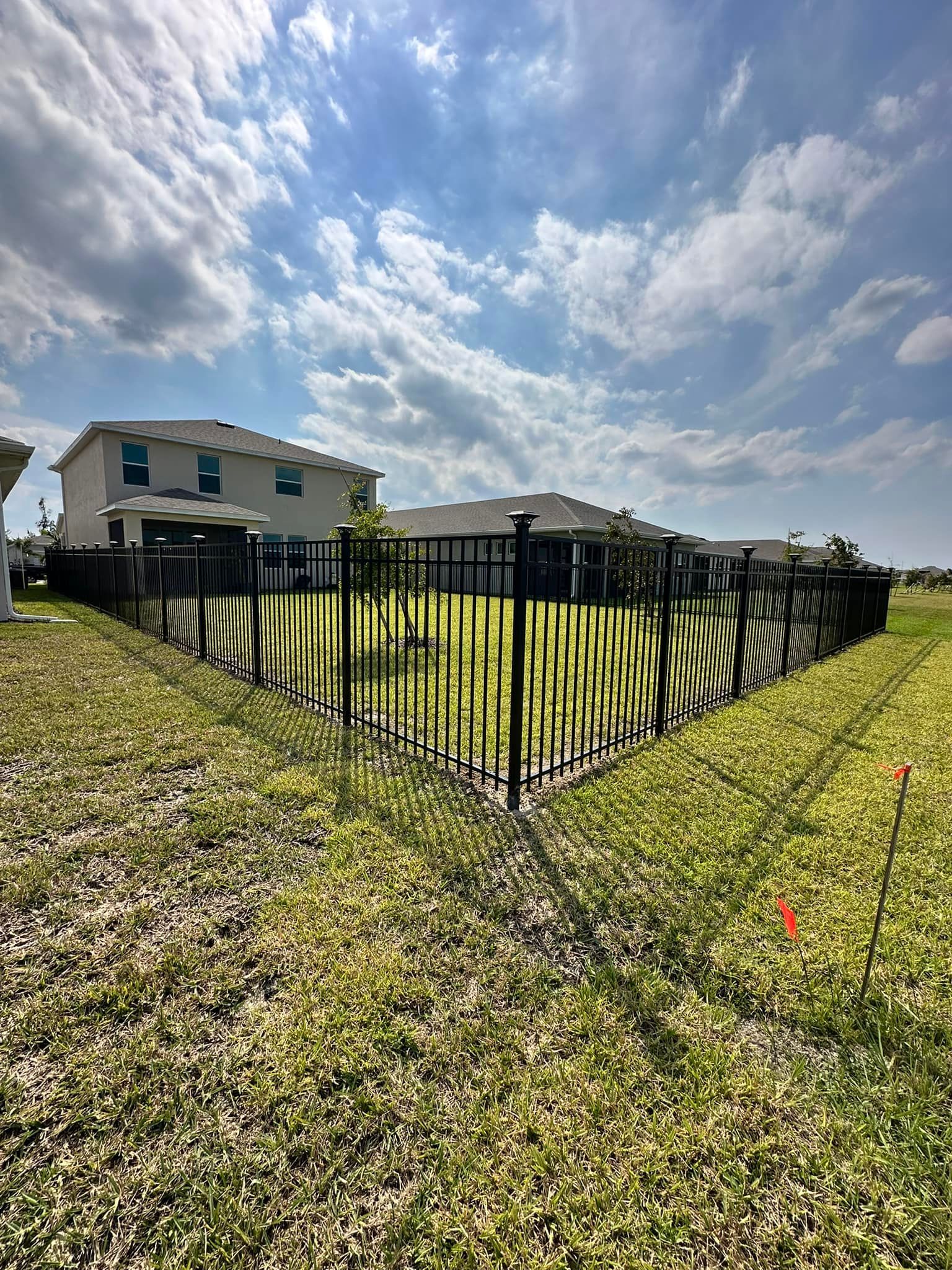 Black fence encloses a backyard with a house under a cloudy blue sky.