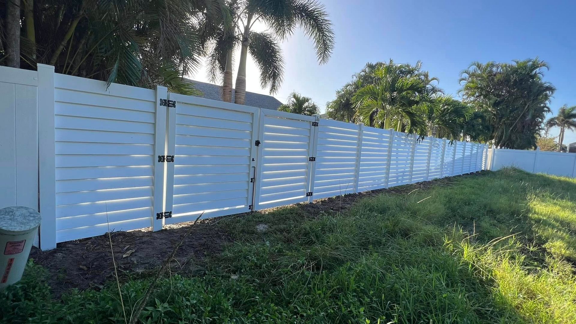 White slatted fence alongside a grassy area; sunny day with trees in the background.