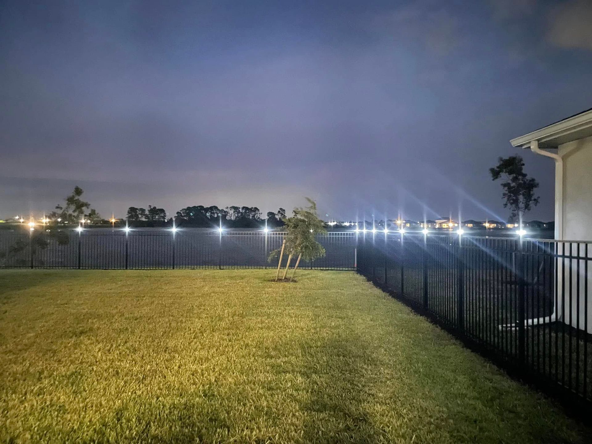 Backyard at night, with a grassy lawn, black fence lined with lights, and a cloudy sky.