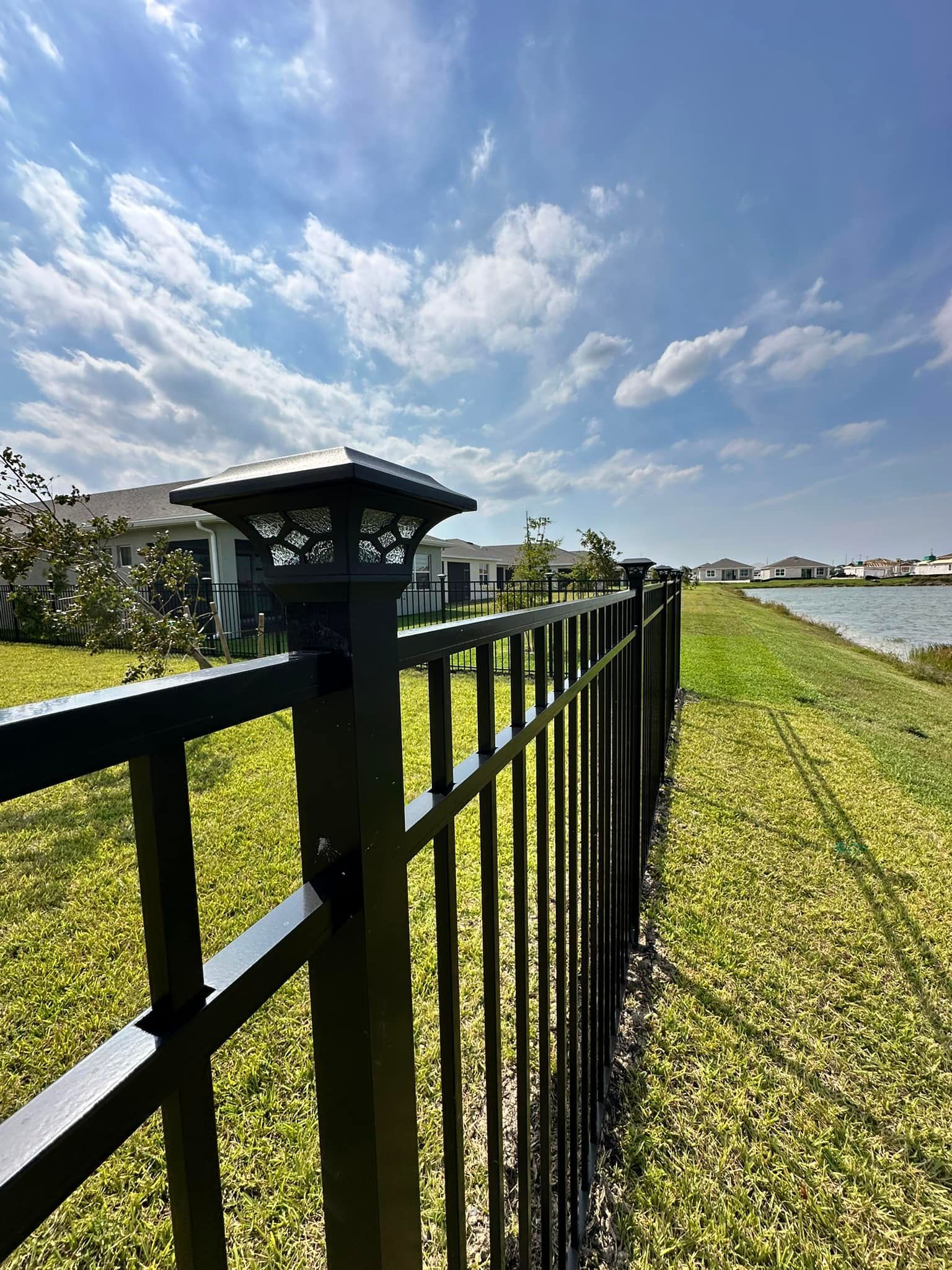 Black fence with solar lights along grassy waterfront, blue sky.